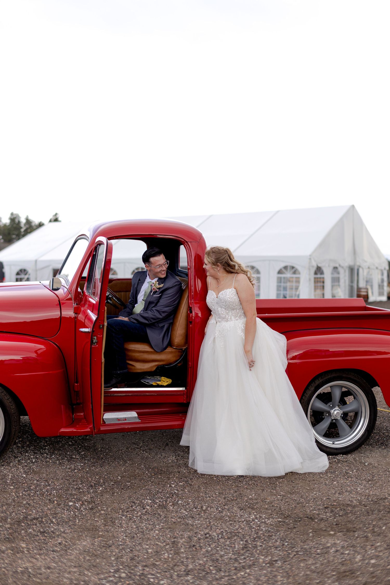 Bride in white tulle gown admires groom seated in classic red vintage pickup truck at outdoor wedding venue.