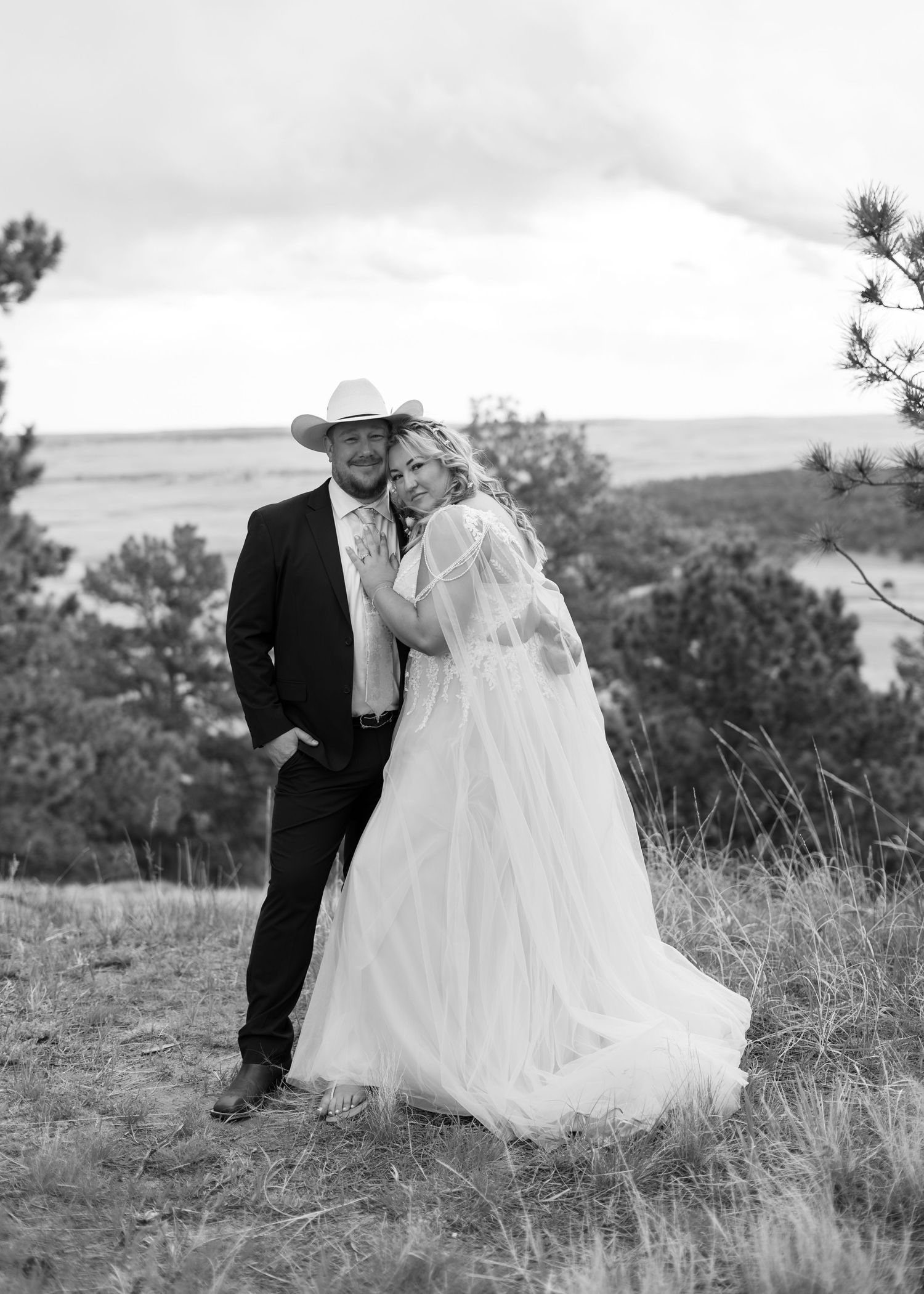 Black and white wedding photo of a couple in a field, groom in cowboy hat and suit, bride in flowing white gown.