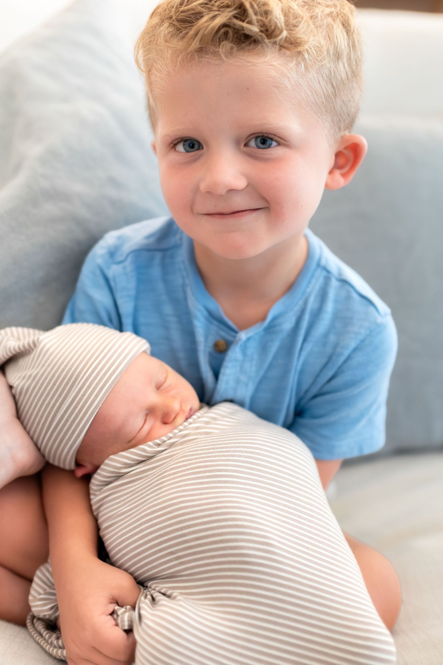 Young boy with blue eyes smiles while holding a sleeping swaddled newborn baby in striped wrap.