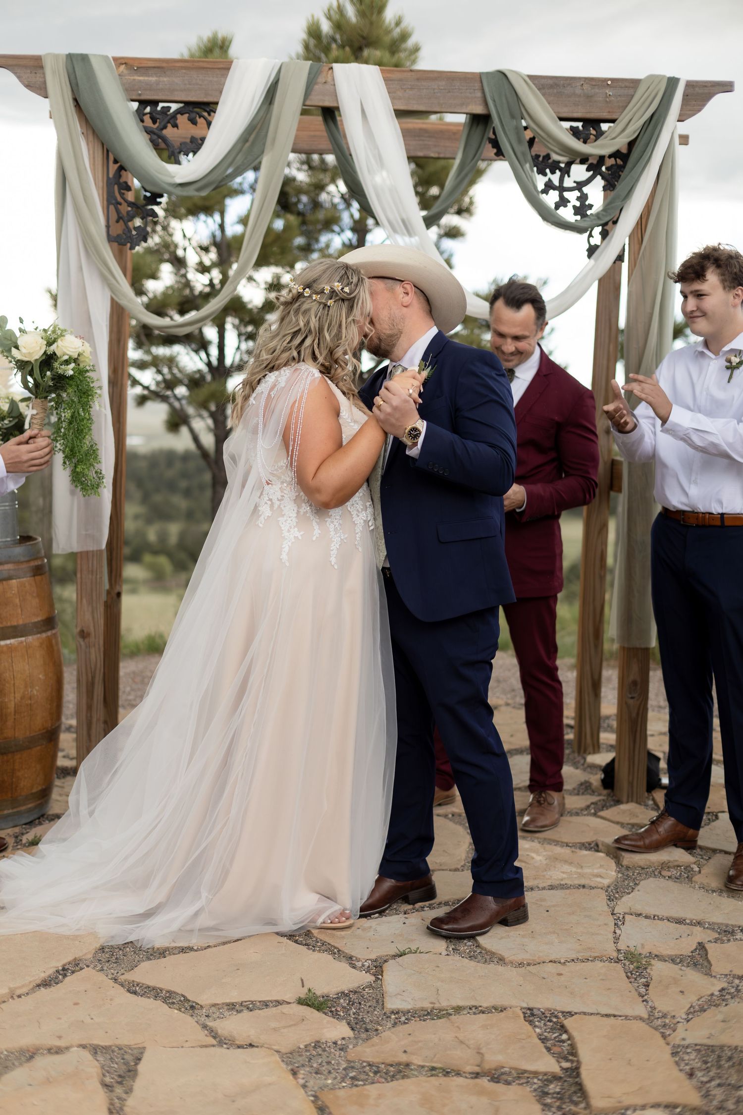 Bride and groom share first kiss at rustic outdoor wedding ceremony under wooden arch with sage green draping.