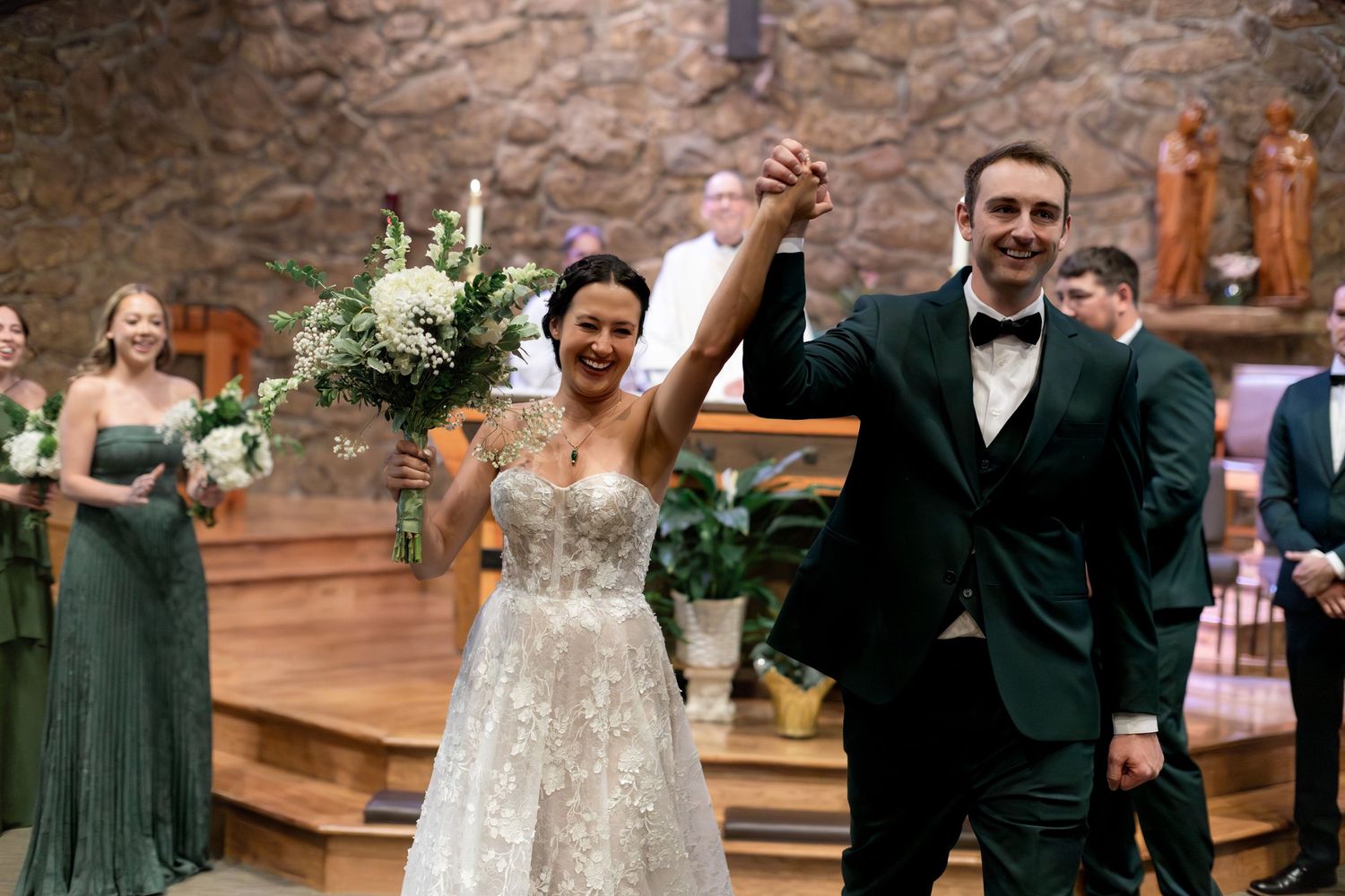 Joyful newlyweds raising hands at church altar, bride in lace gown holding bouquet, groom in green tuxedo.