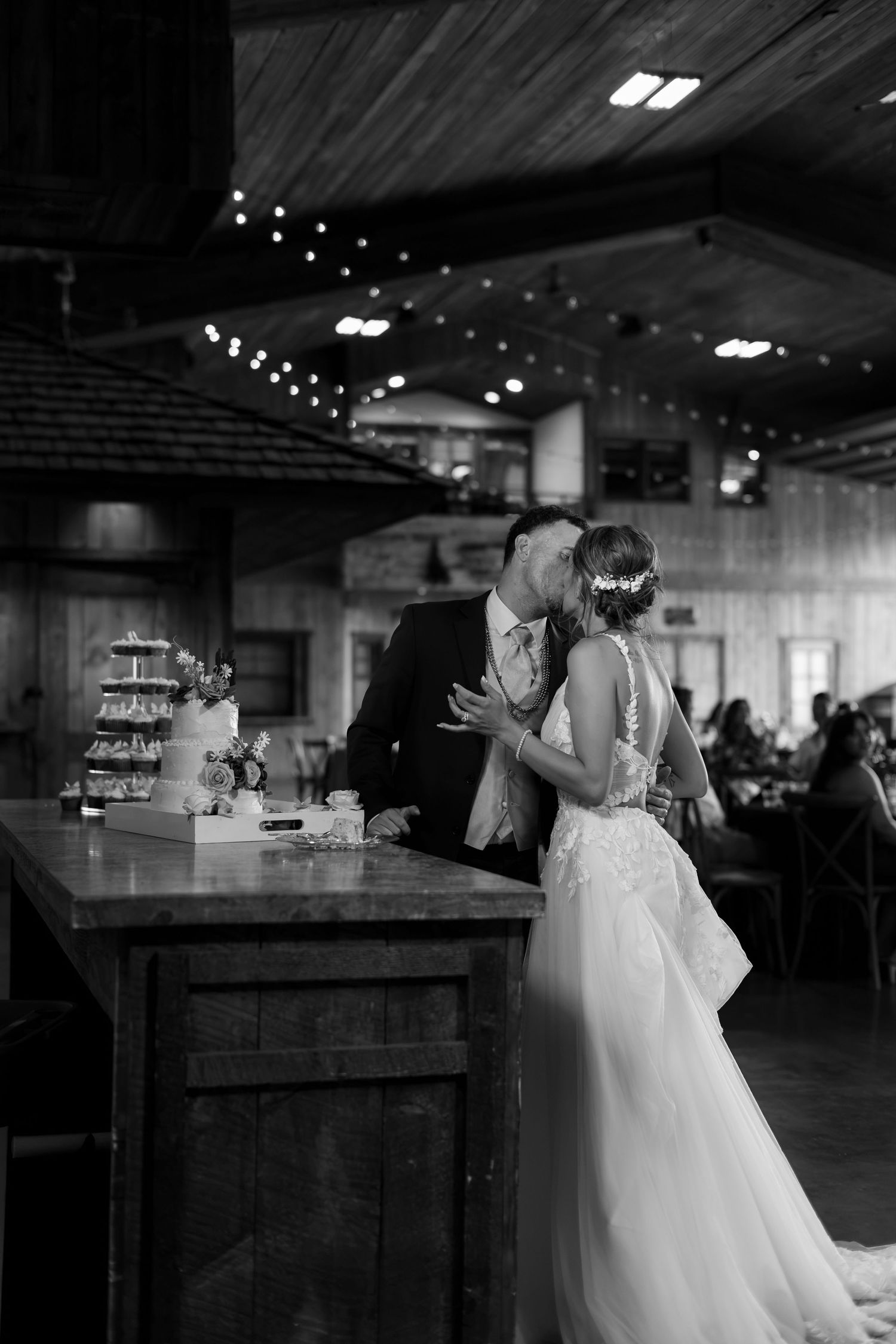 Black and white photo of newlyweds sharing kiss while cutting wedding cake at rustic barn reception.