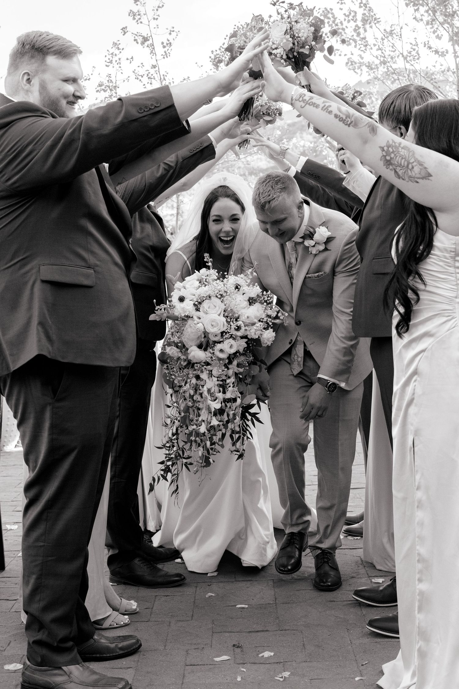 Black and white photo of a joyful bride and groom walking under an arch of raised hands formed by their wedding party.