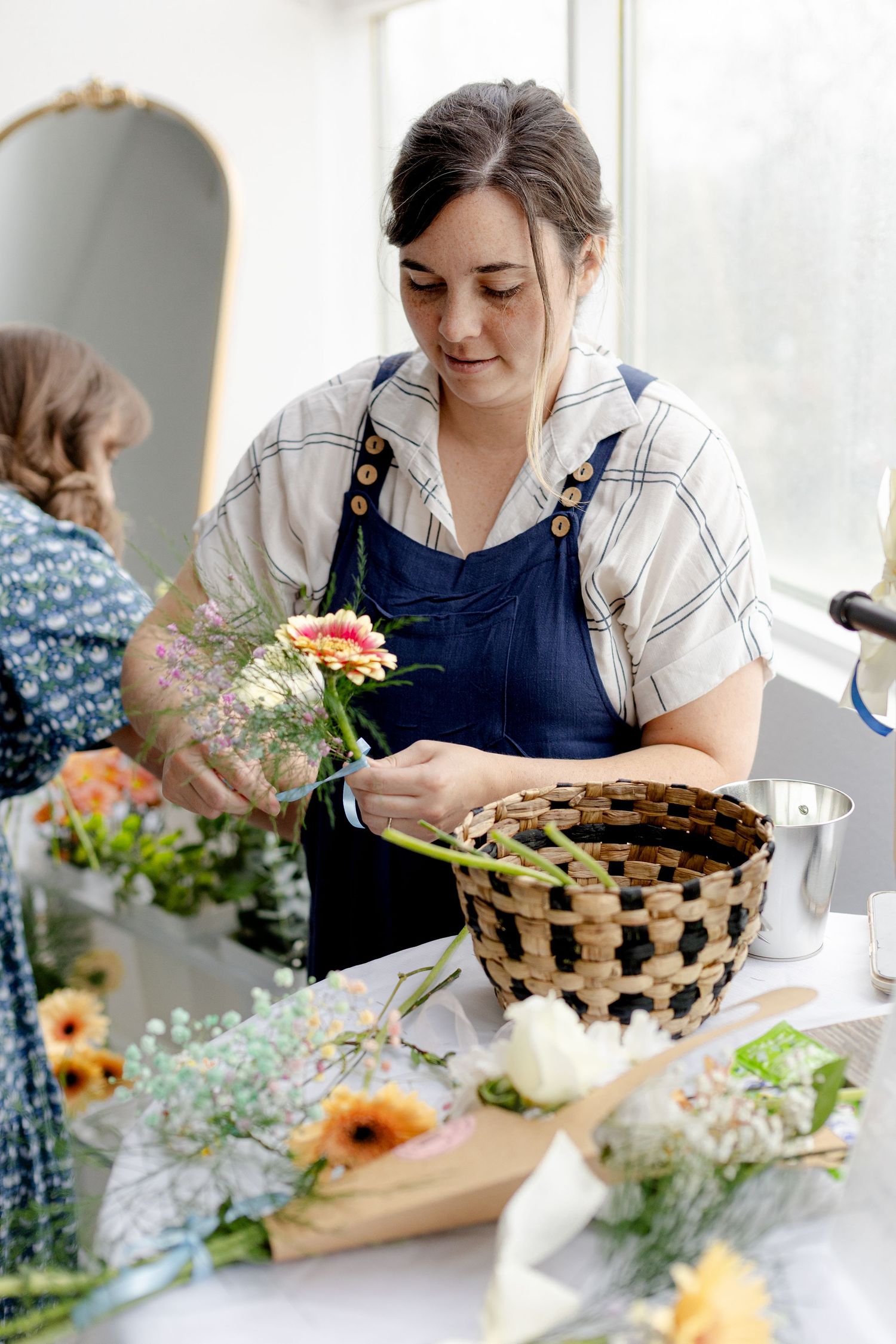 Woman in blue apron arranging colorful flowers into a woven basket at a floral design workshop.
