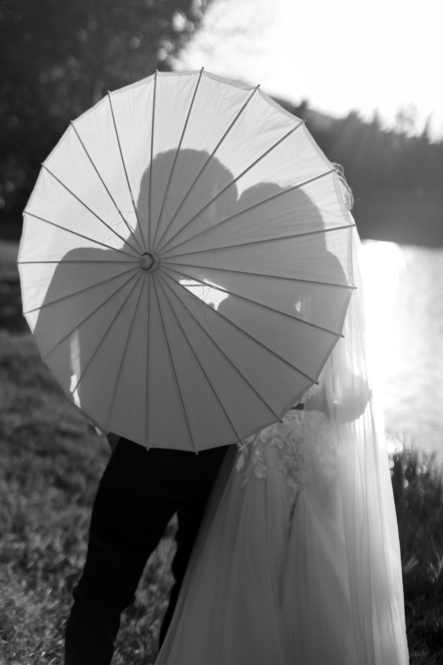 Black and white photo of bride and groom hiding behind a parasol, casting a romantic heart-shaped shadow by a lake.