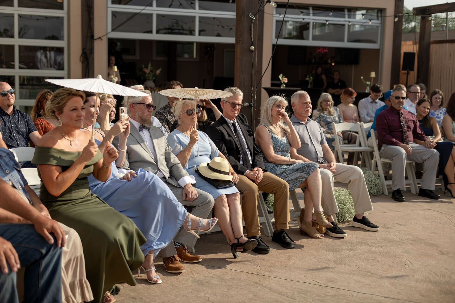 Wedding guests seated outdoors in sunshine, some shielding from sun with hats during ceremony.