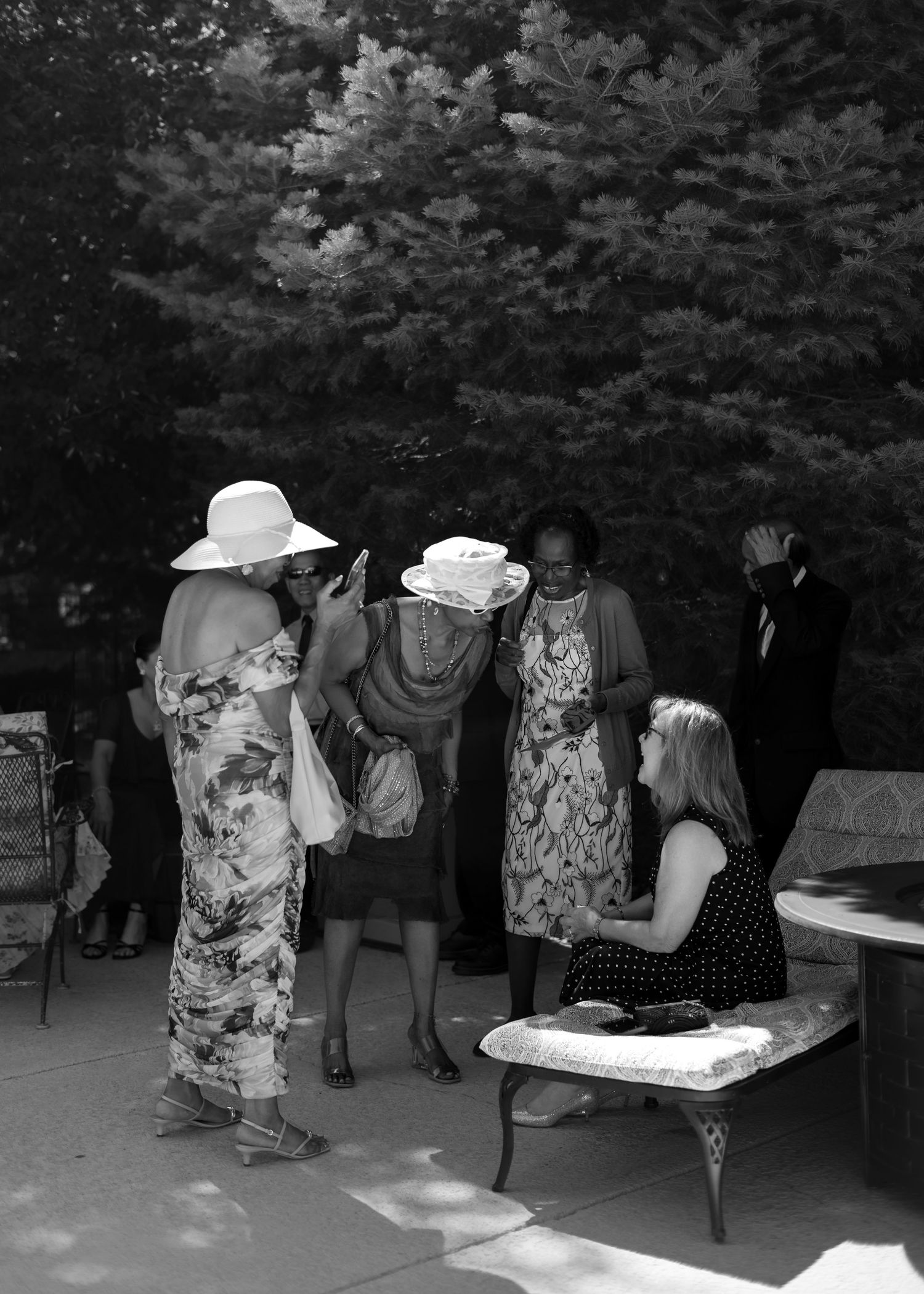 Elegantly dressed wedding guests in wide-brim hats socializing outdoors in black and white.