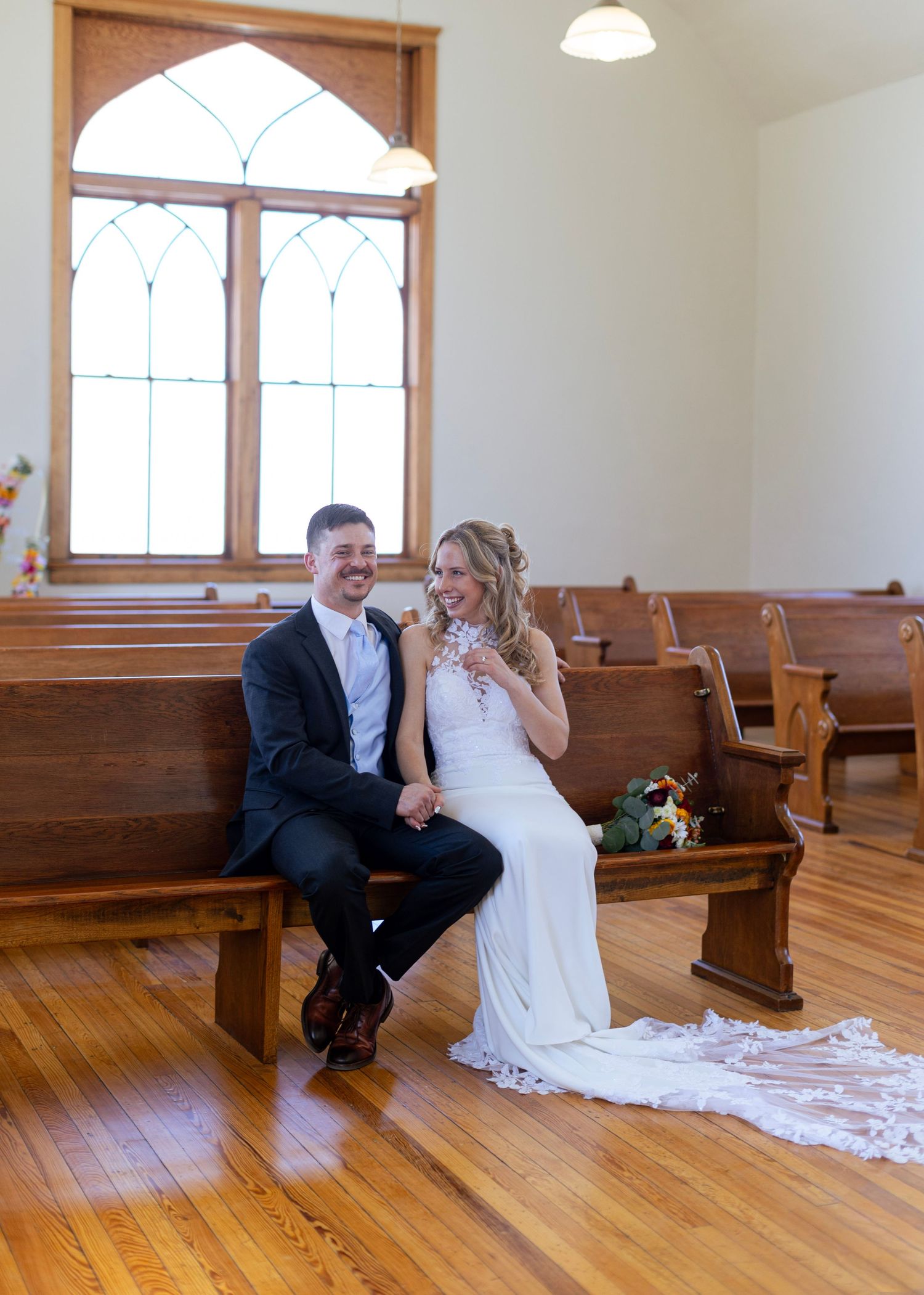 Bride and groom sitting together on a wooden church pew laughing inside a charming chapel.