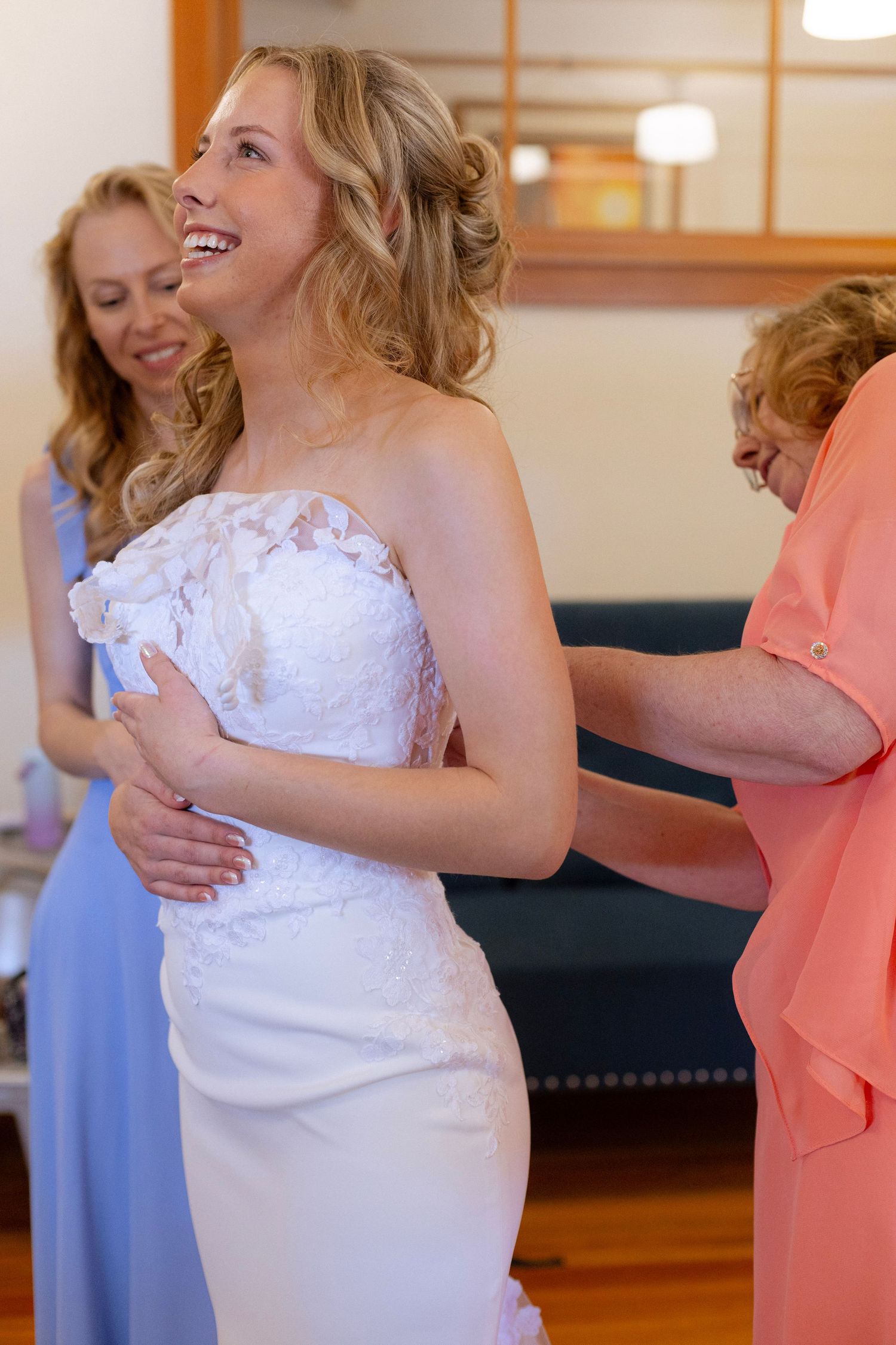 Smiling bride in white lace strapless gown being helped by two women in a bright room.