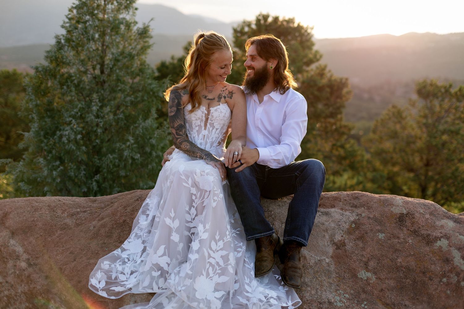 Couple in wedding attire sitting on a rock at sunset with mountain views, bride in lace gown, groom in white shirt.