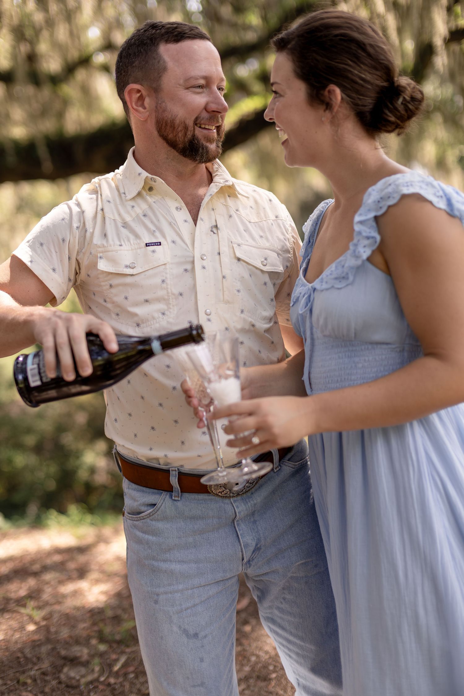 Smiling couple celebrates engagement pouring champagne together in a sunlit wooded setting.
