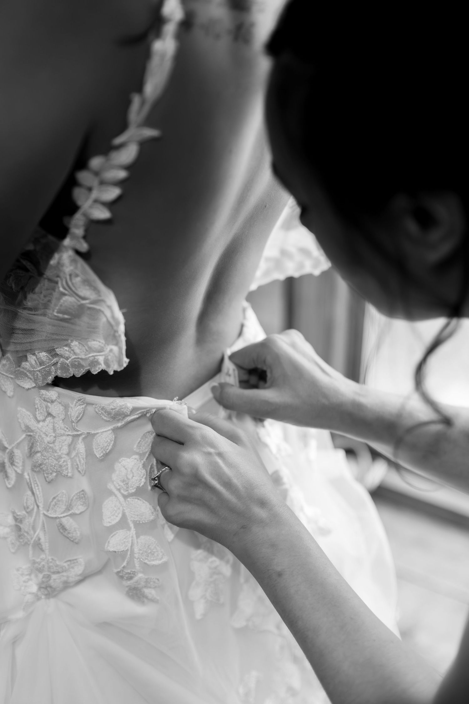 Bridesmaid carefully fastening buttons on intricate lace wedding gown during bridal preparations.