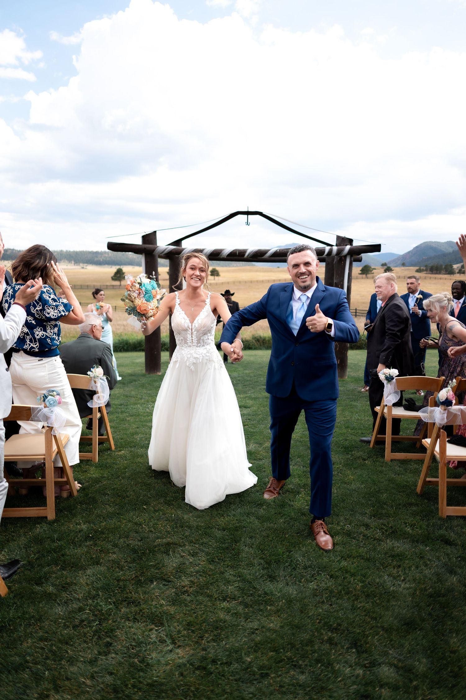 Bride and groom walk down outdoor aisle holding hands after wedding ceremony with mountain backdrop.