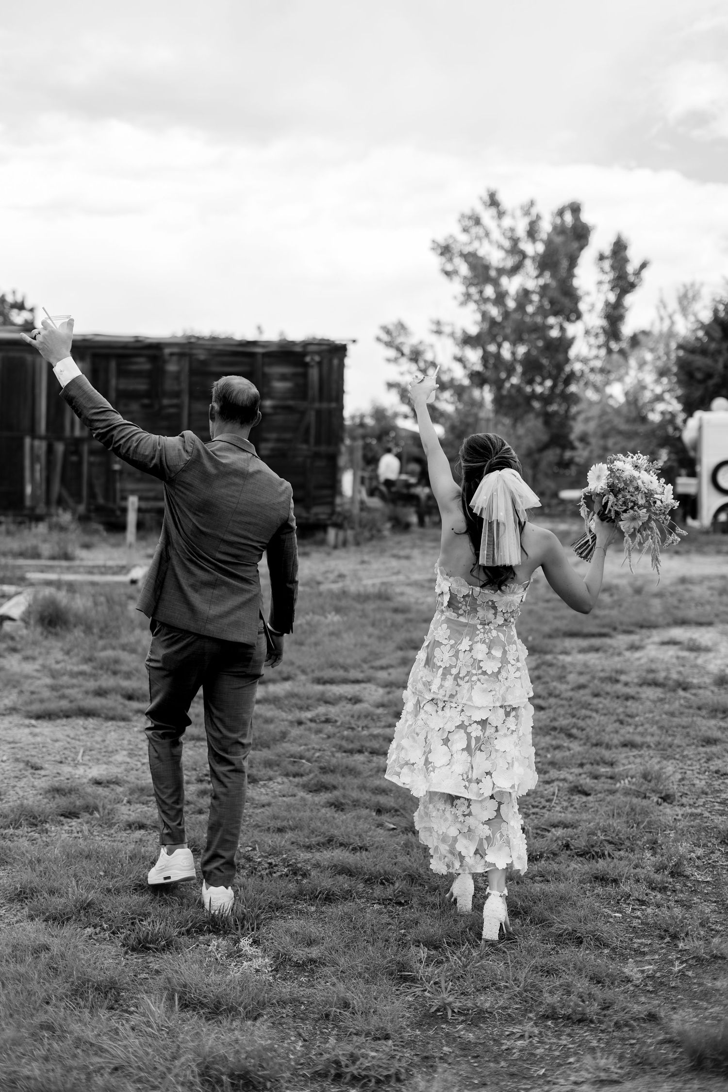 Black and white photo of a bride and groom walking away, arms raised in celebration, bride holding bouquet.