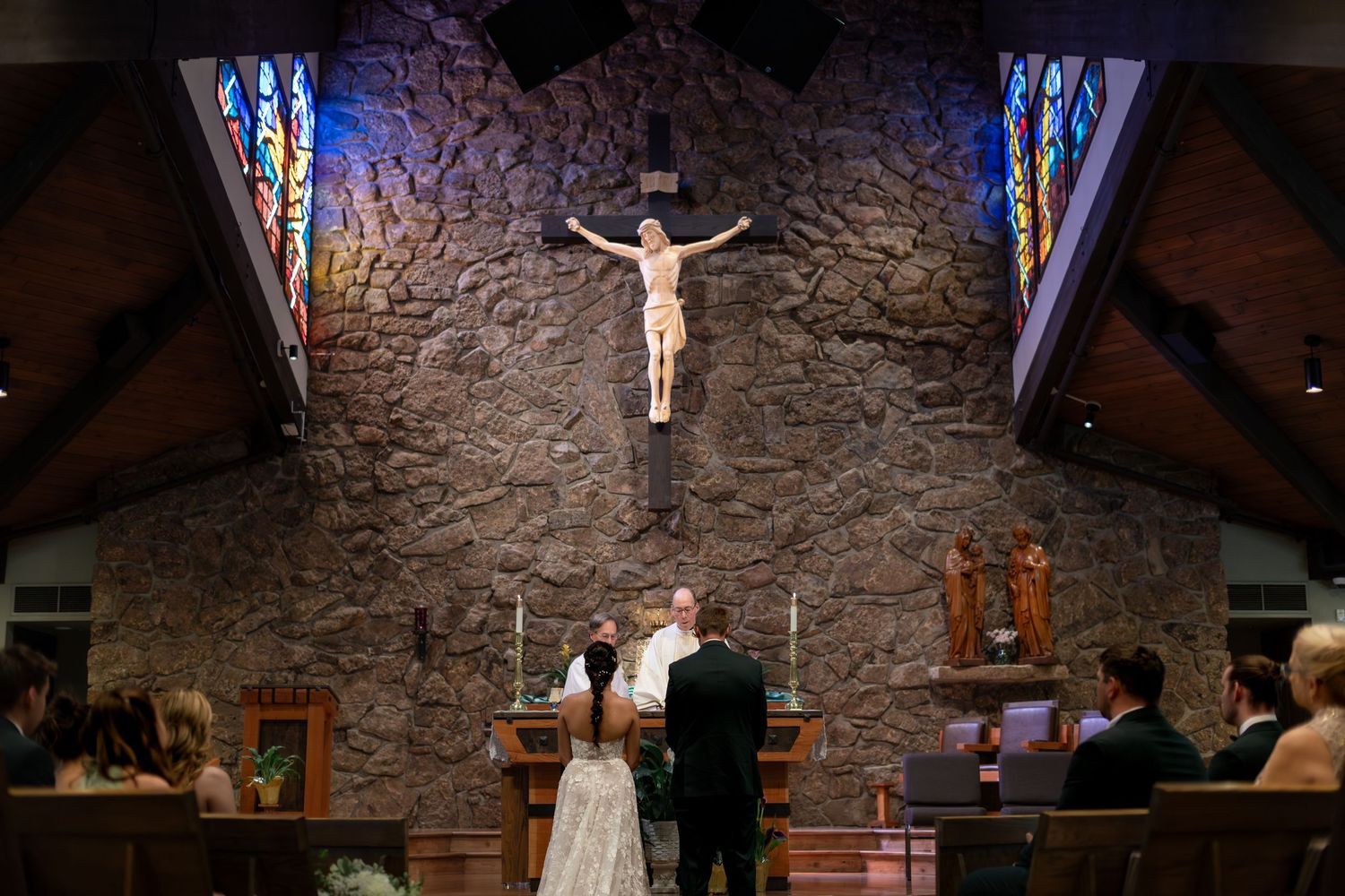 Catholic church wedding ceremony with bride and groom at altar beneath crucifix and stained glass windows.