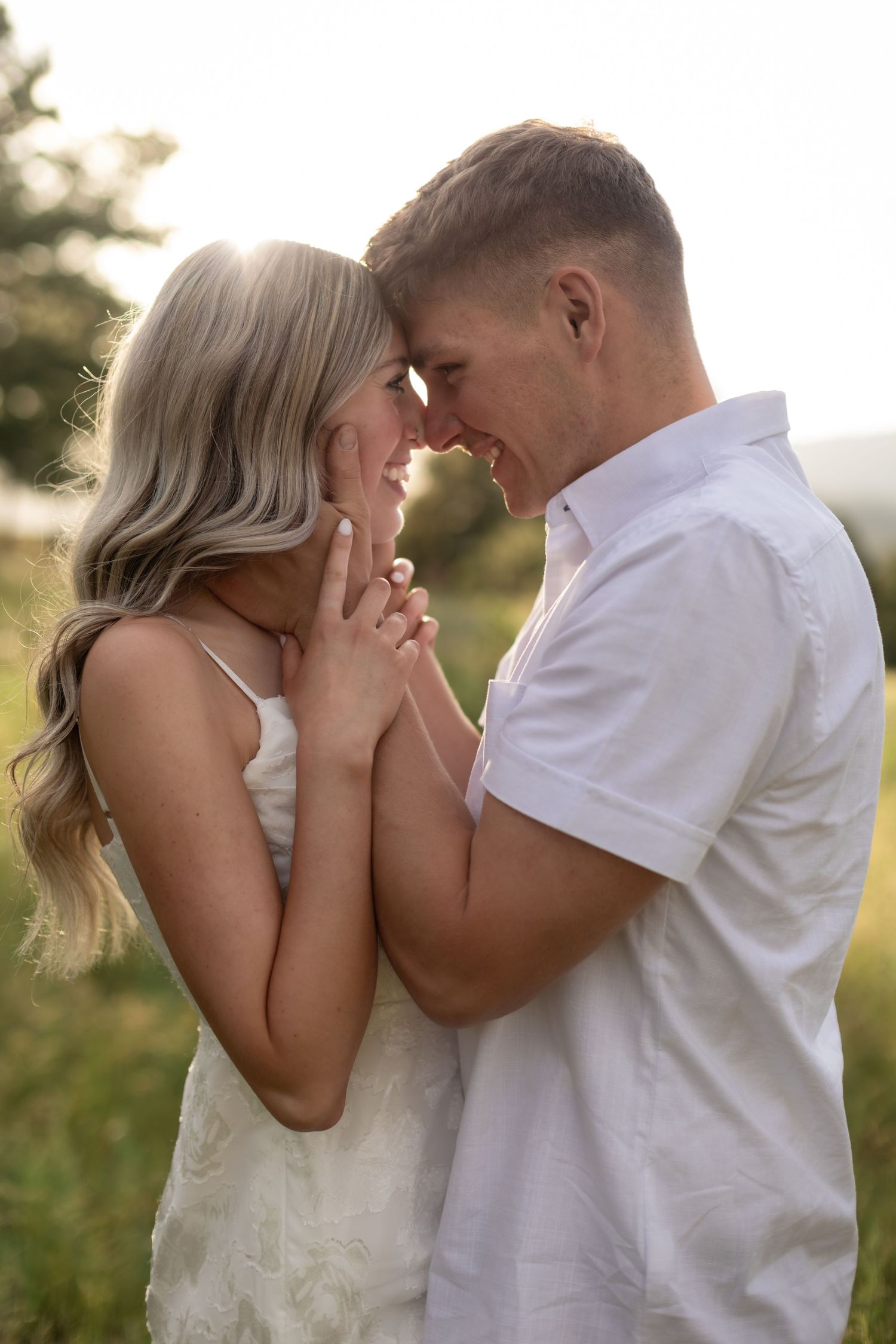 A romantic couple in white outfits touching foreheads in a sunlit field, sharing an intimate moment.