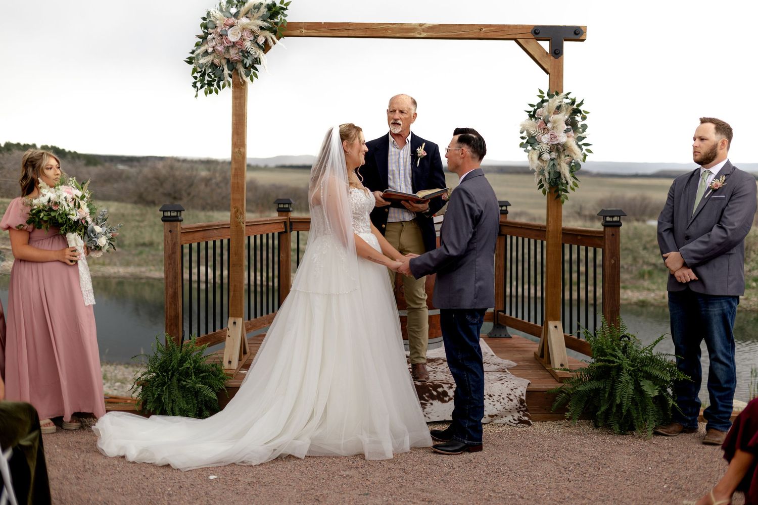 Outdoor wedding ceremony under wooden arch with floral decor, bride in white gown, groom in gray suit, officiant, lakeside backdrop.