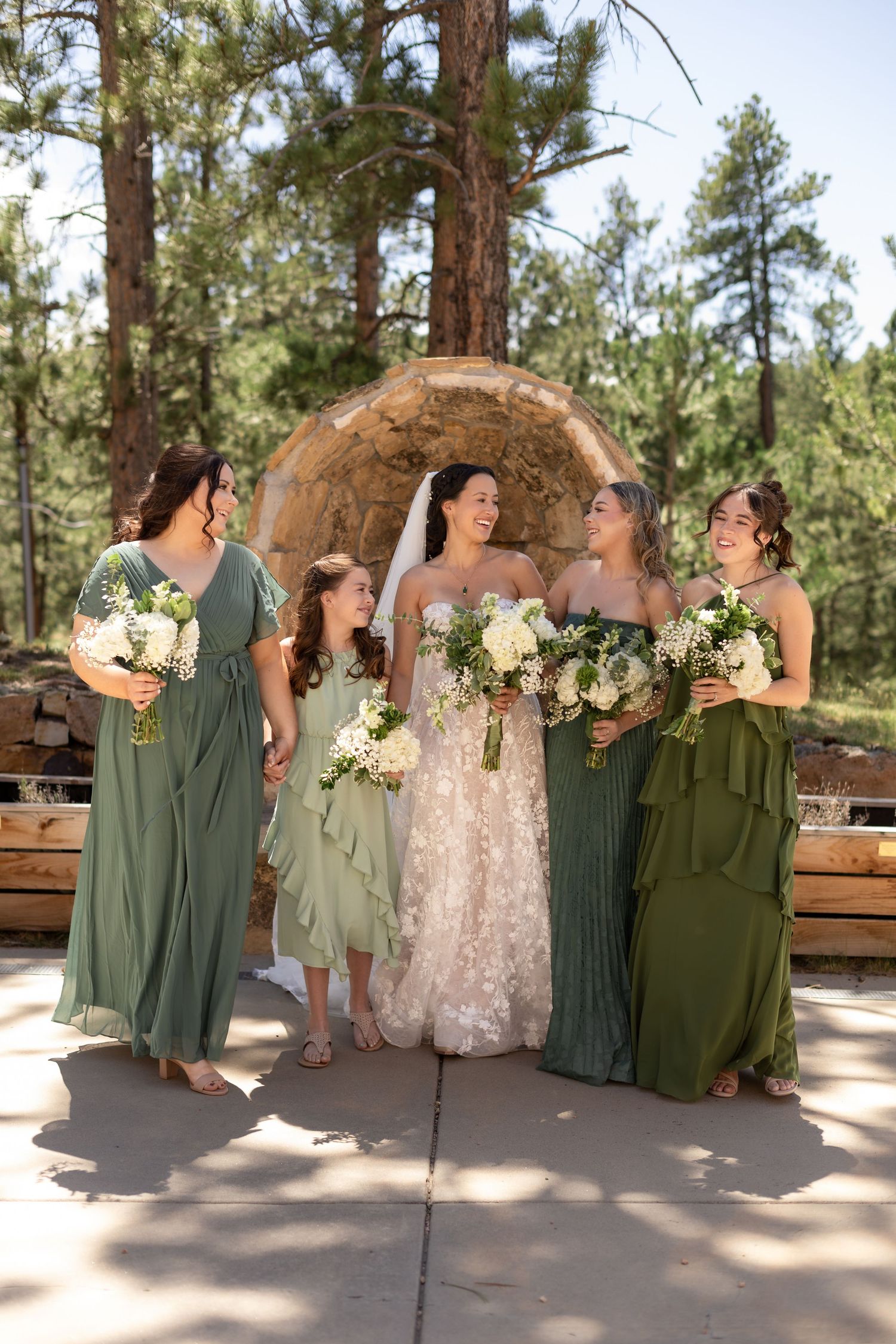 Bride in white gown with bridesmaids in sage green dresses holding white bouquets at outdoor forest wedding ceremony.