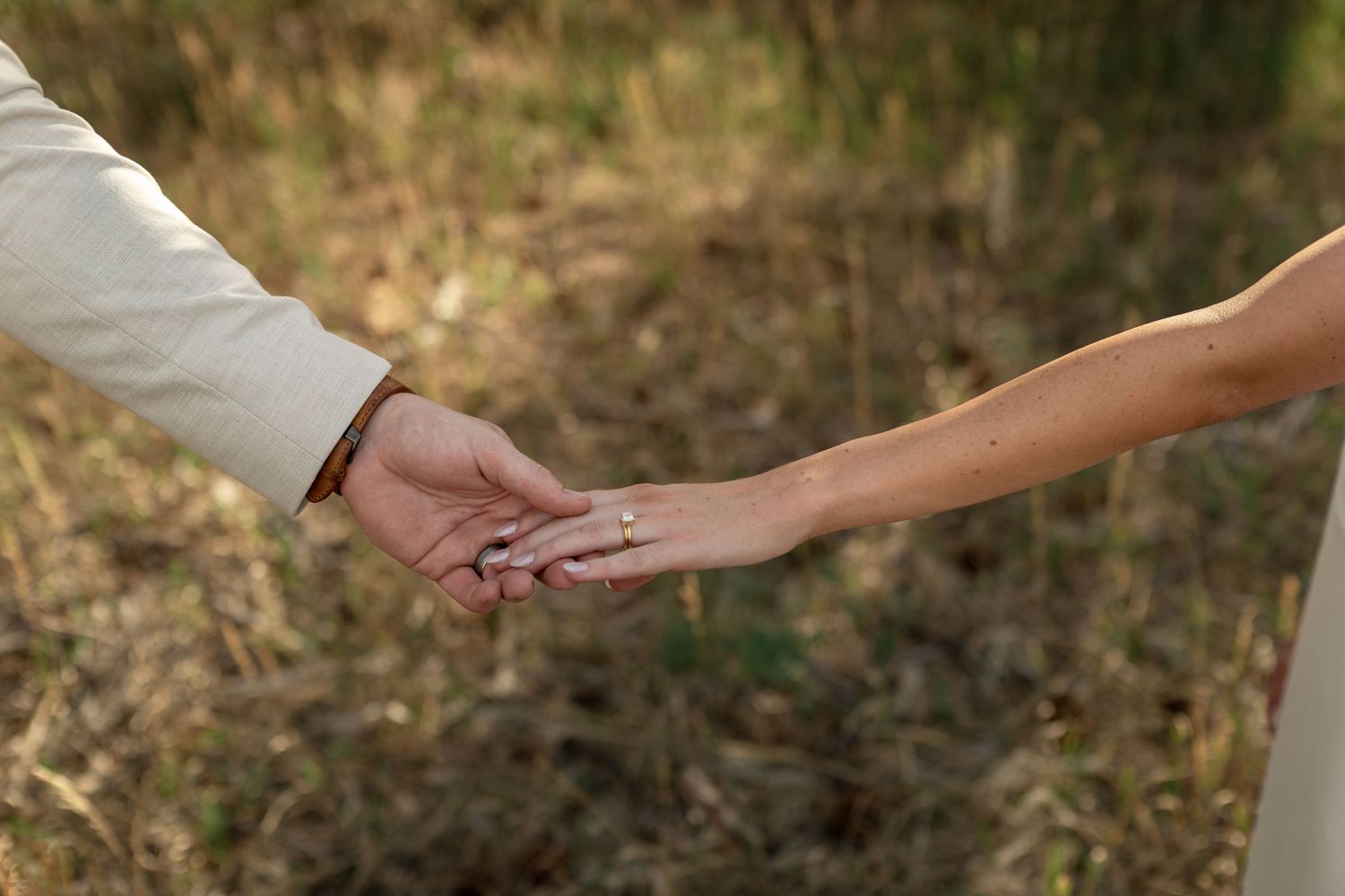 Couple holding hands outdoors in a field, woman wearing an engagement ring, romantic wedding photo.