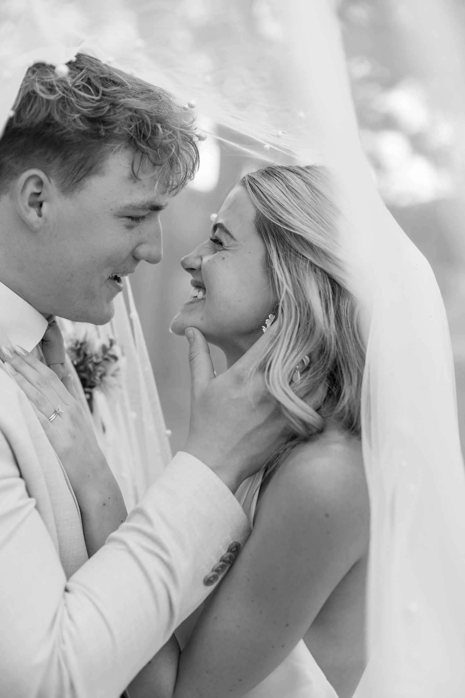 Black and white photo of a smiling bride and groom sharing an intimate moment under a veil on their wedding day.