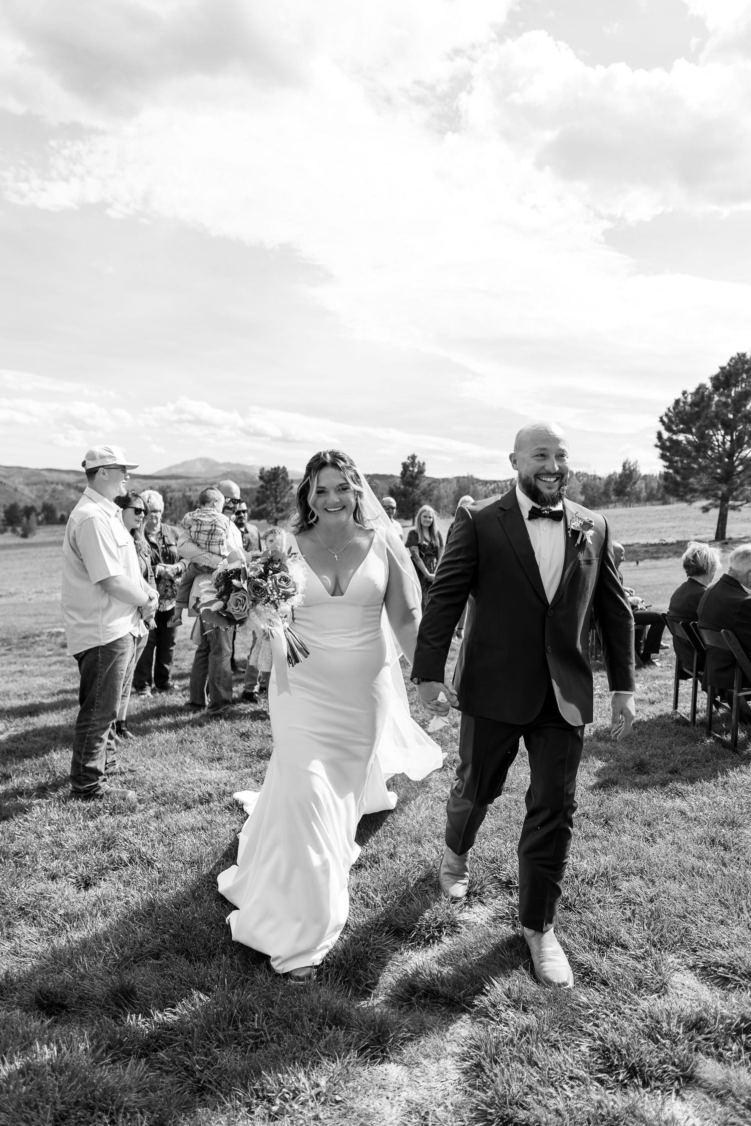Joyful newlyweds walk back down aisle after outdoor wedding ceremony surrounded by guests in black and white photo.