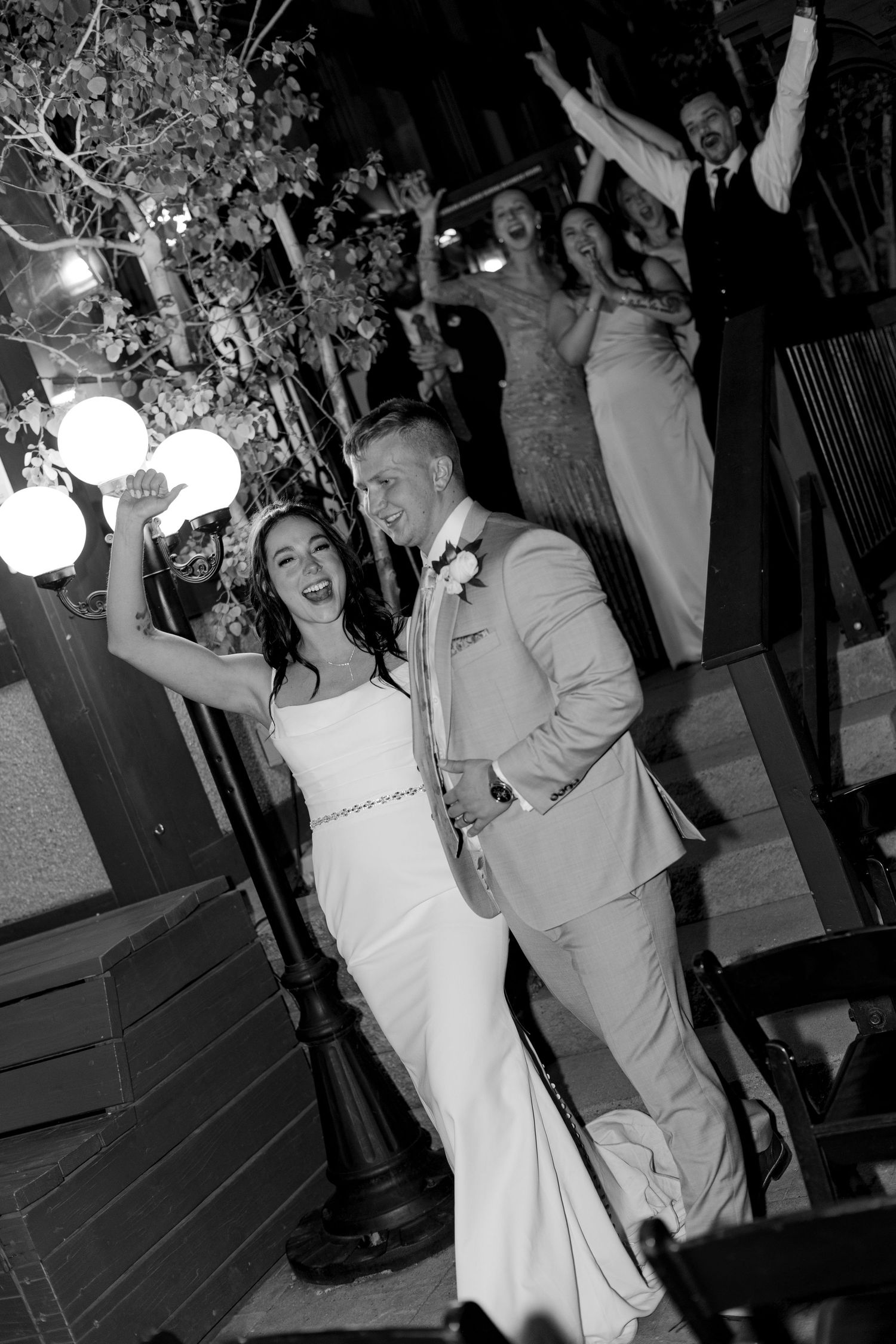 Black and white photo of joyful bride and groom grand entrance with cheering wedding party behind them.