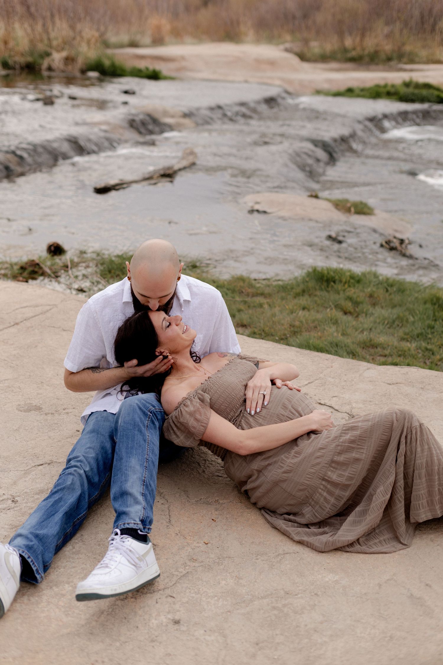 Couple sharing a tender moment outdoors, pregnant woman resting on man's lap near a rocky riverbed.