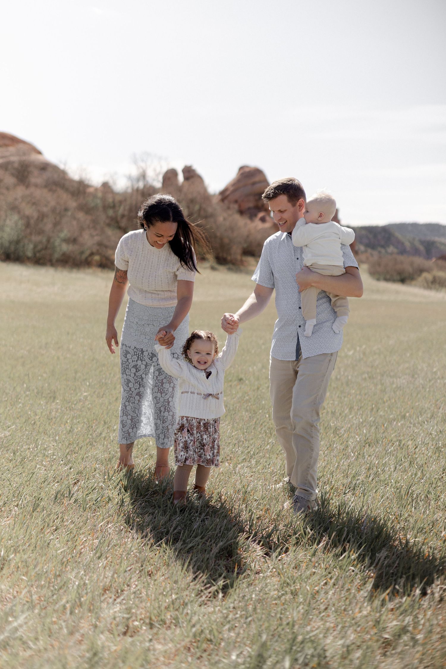 Family of four walking in a sunny field with red rock formations, parents holding children's hands outdoors.