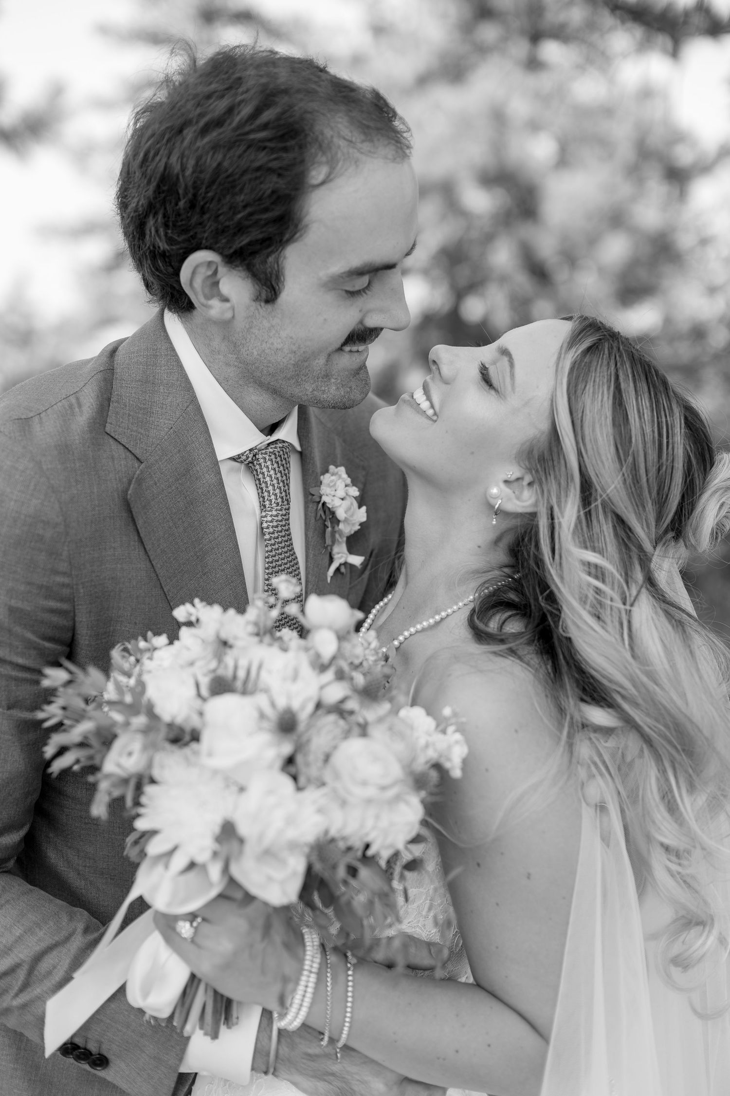 Black and white wedding photo of smiling bride holding bouquet and groom leaning in for a kiss outdoors.