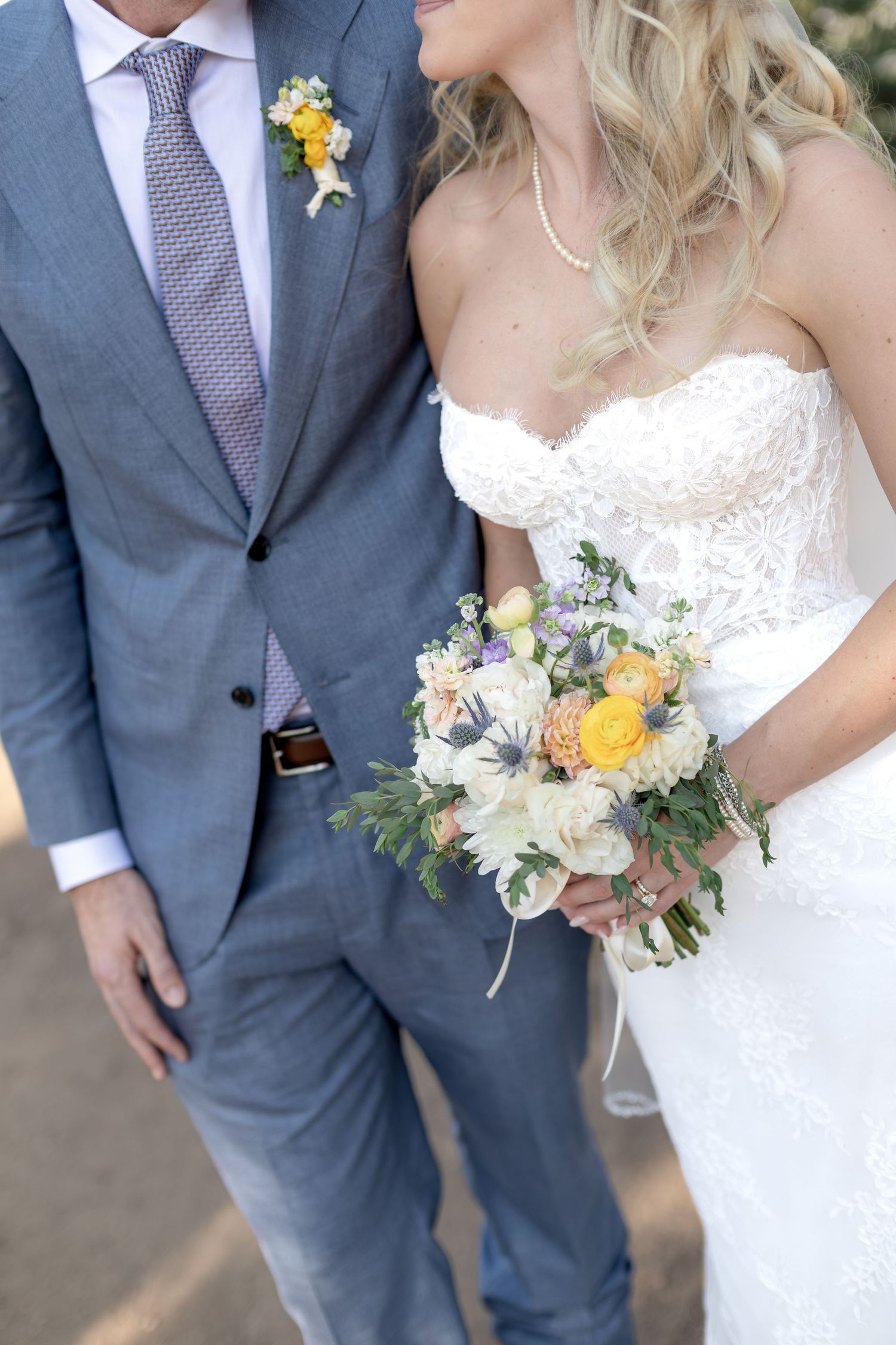 Bride and groom posing together, bride holding colorful bouquet with yellow, white, and purple flowers in lace strapless gown.