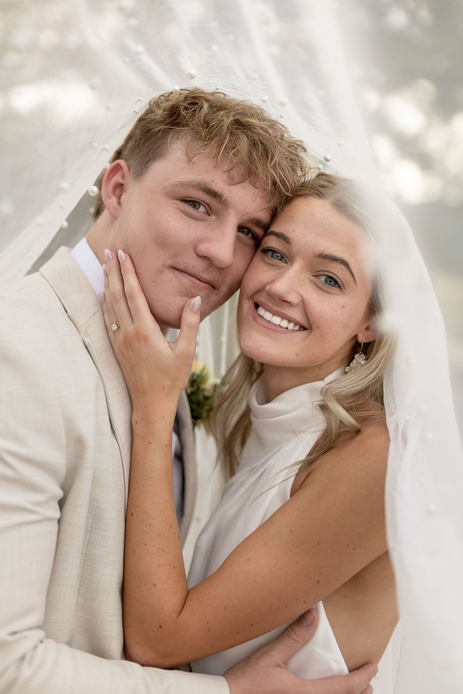 A smiling bride and groom pose closely together under a white veil on their wedding day.