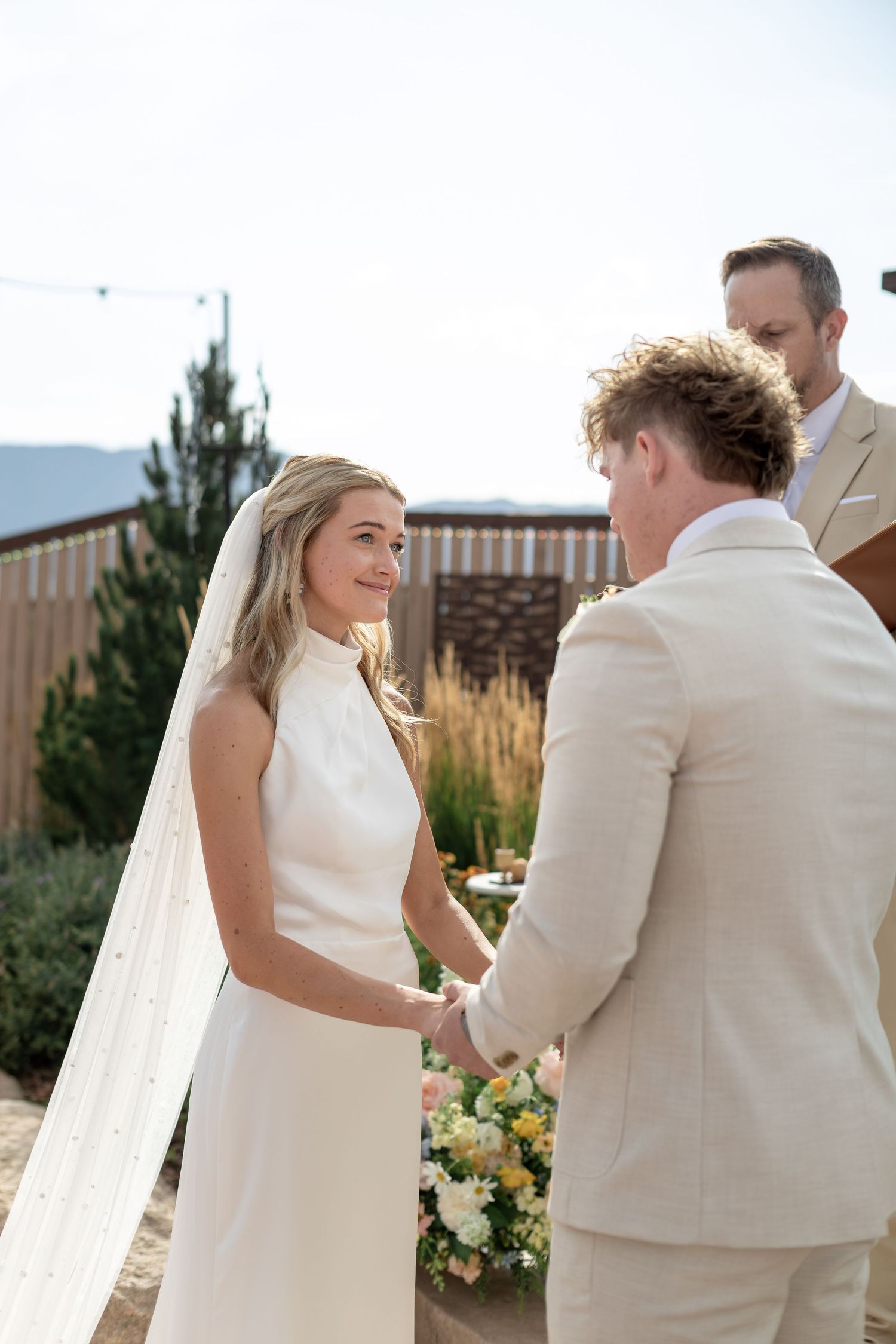 Smiling bride in white halter gown holds grooms hands at sunny outdoor wedding ceremony altar.