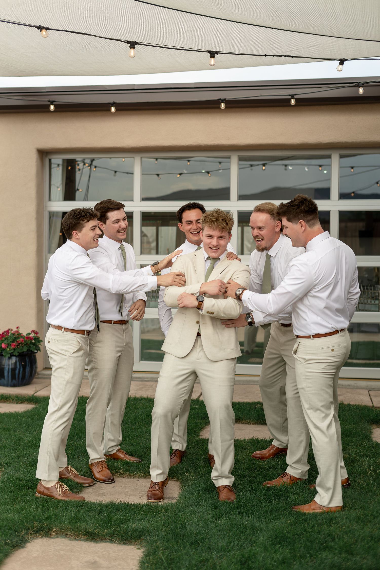 Groom in tan suit surrounded by groomsmen in white shirts and khakis, laughing outdoors at wedding venue.