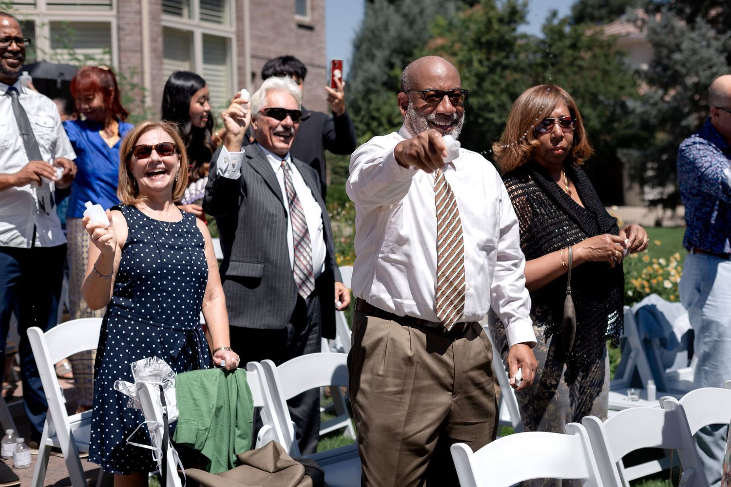 Guests celebrate outdoors at a sunny ceremony, smiling and pointing among white chairs at a formal event.