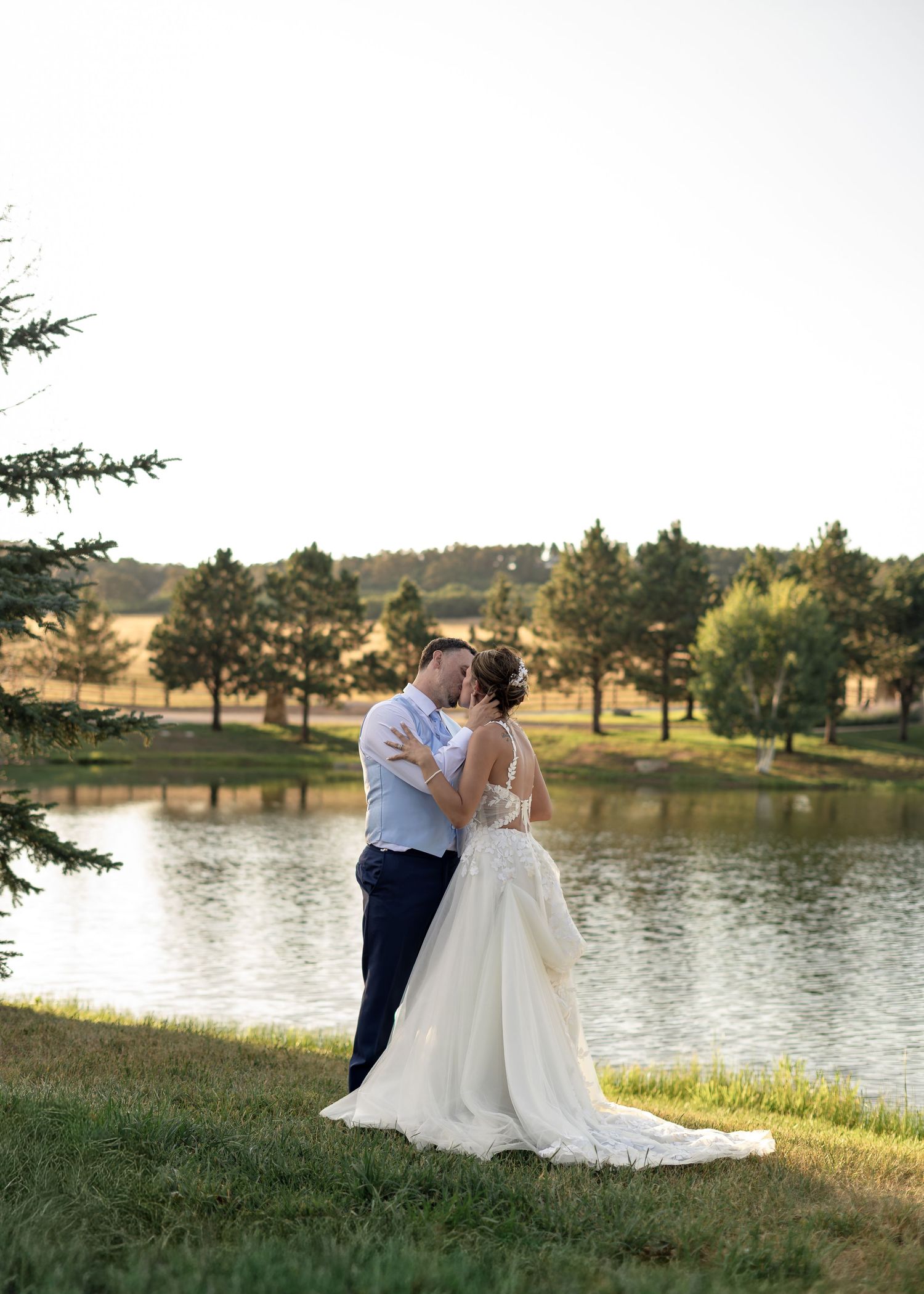 A bride and groom share a romantic kiss by a serene pond surrounded by trees during their outdoor wedding ceremony.
