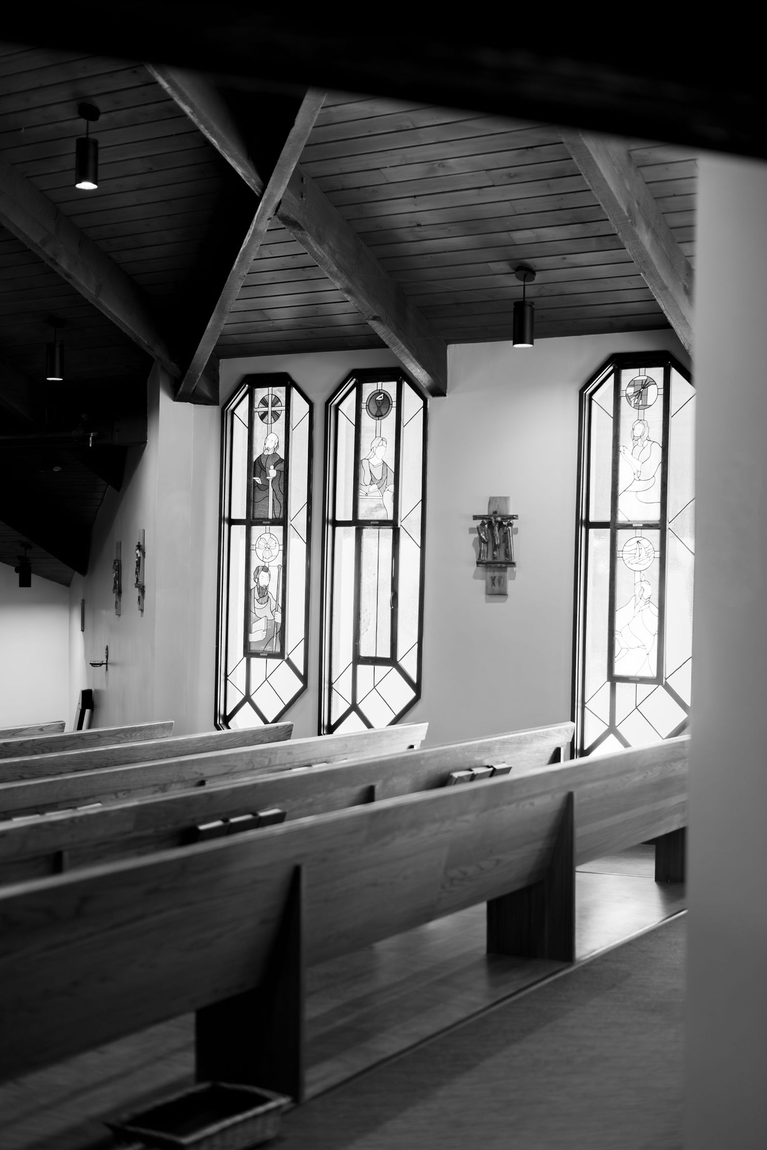 Black and white photo of wooden church pews with stained glass windows and vaulted ceiling interior.