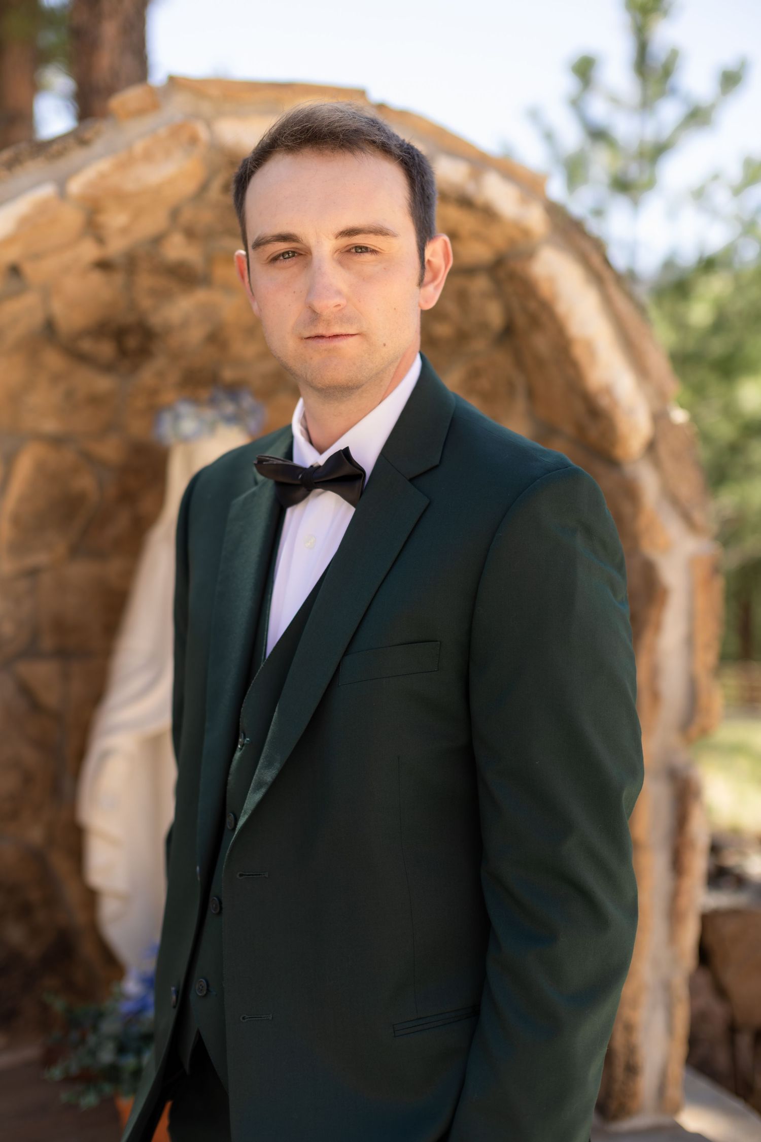 Groom in dark green tuxedo with black bow tie posing in front of stone arch at outdoor wedding venue.