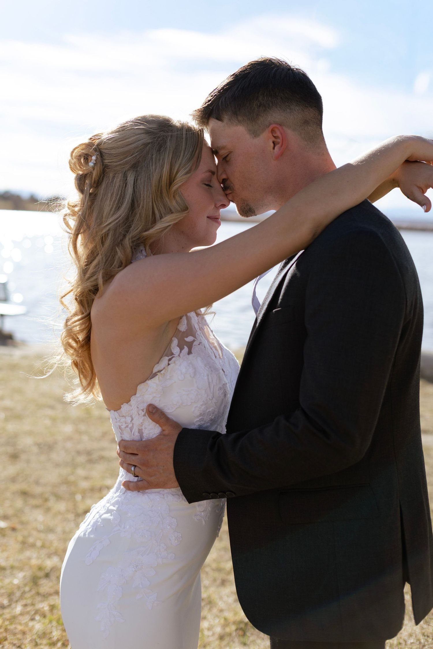 Bride and groom sharing a romantic almost-kiss embrace outdoors near a shimmering lake on their wedding day.