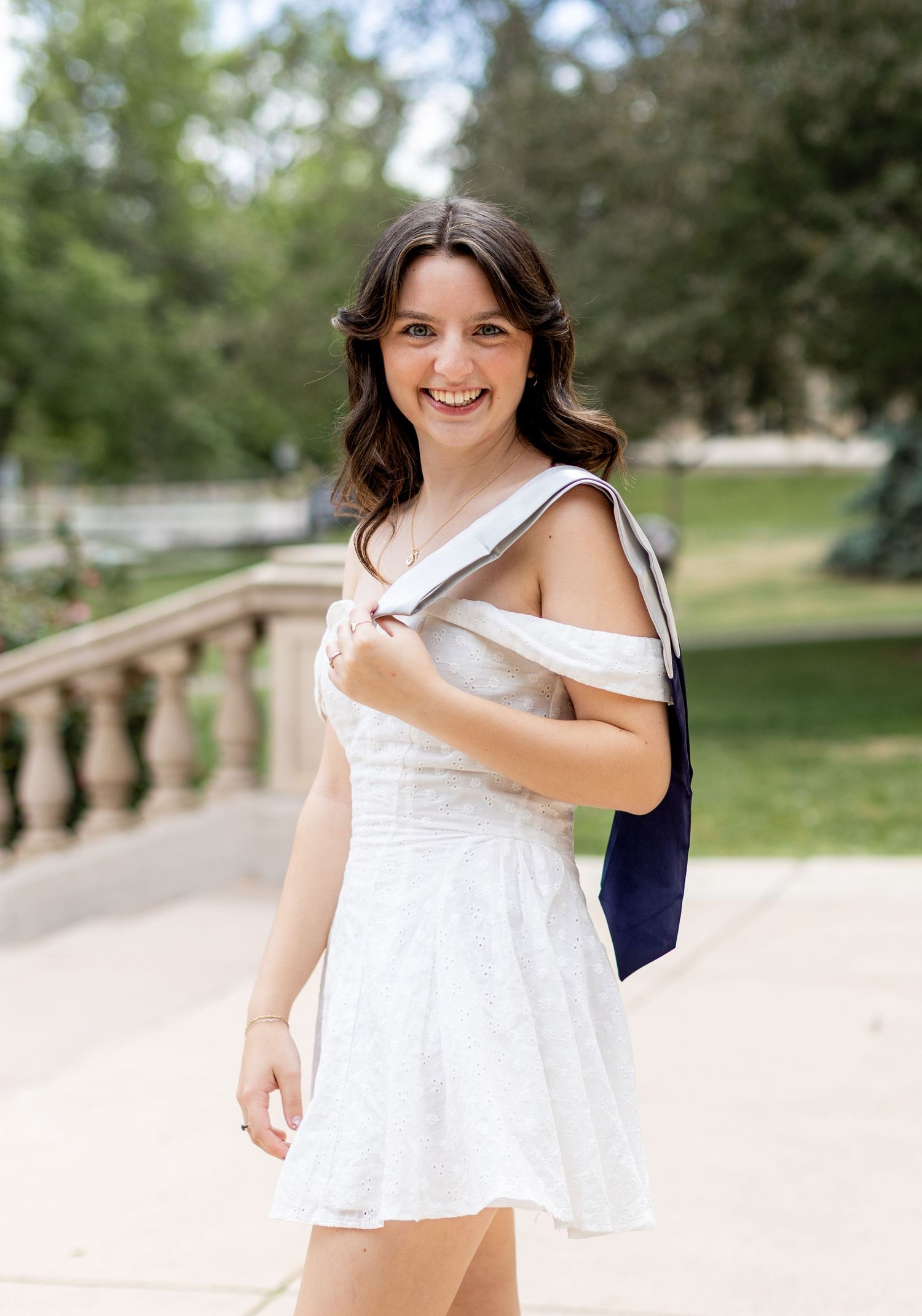 Smiling young woman in white off-shoulder dress carrying navy bag outdoors near stone bridge.
