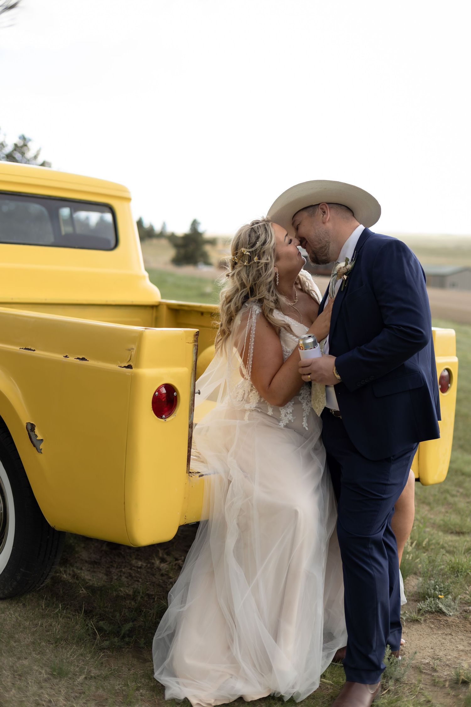 A bride and groom share a kiss beside a vintage yellow pickup truck in a rustic outdoor wedding setting.