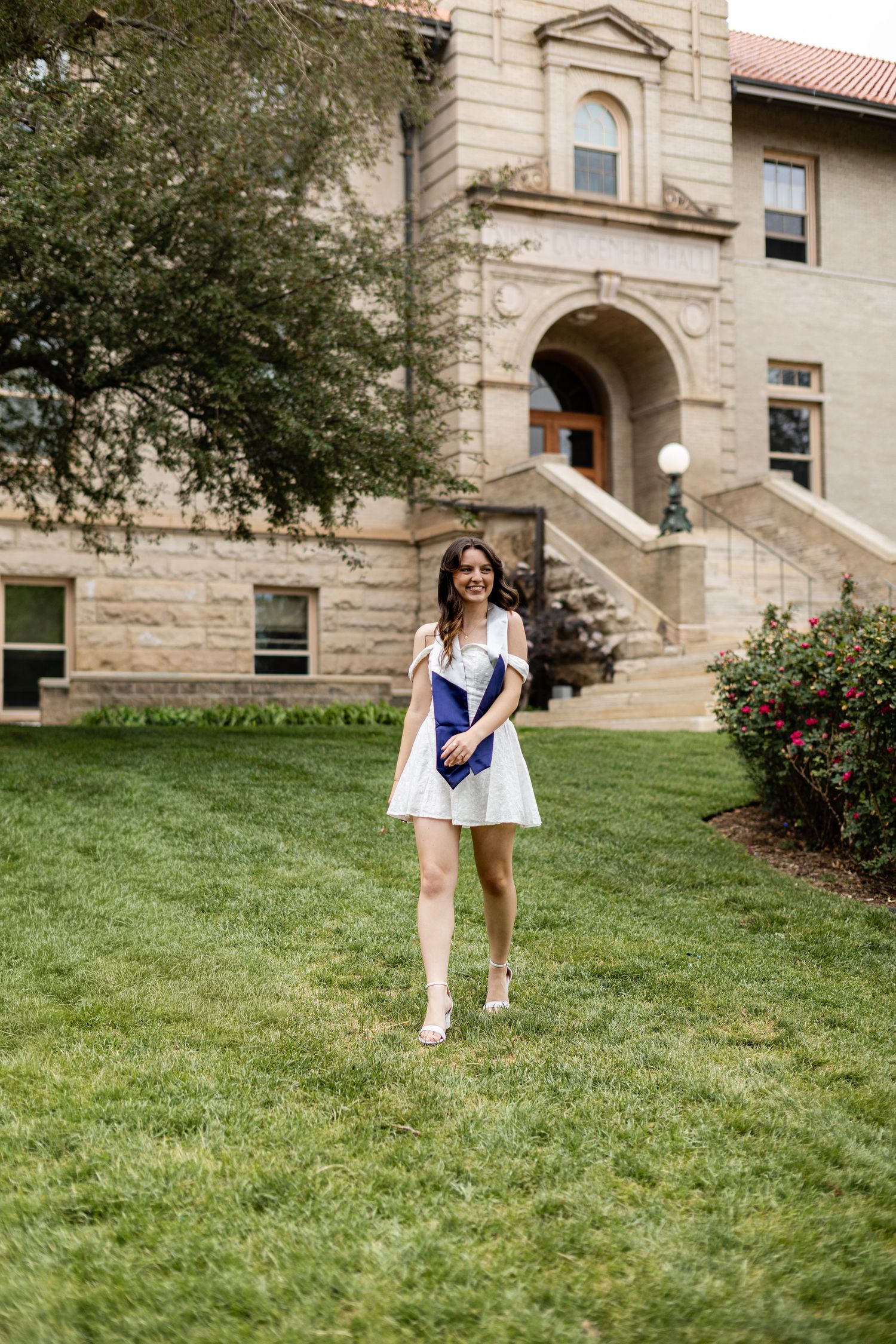 Happy female graduate in white dress holding diploma walking on green campus lawn.