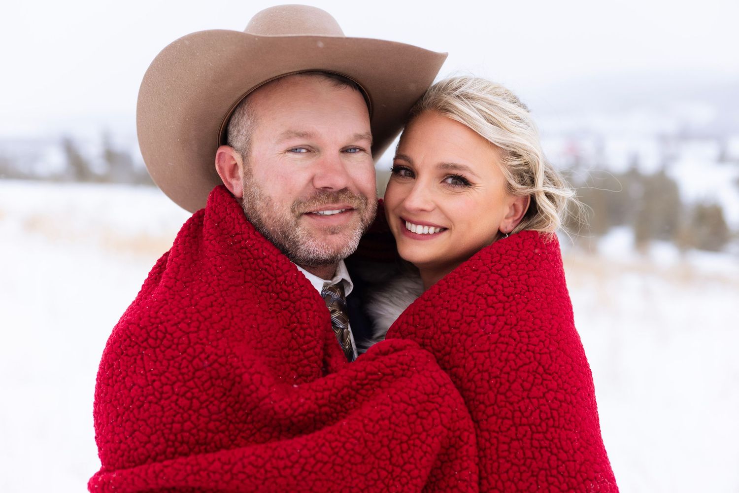 A couple wrapped in a red blanket share an intimate moment in a snowy winter landscape.