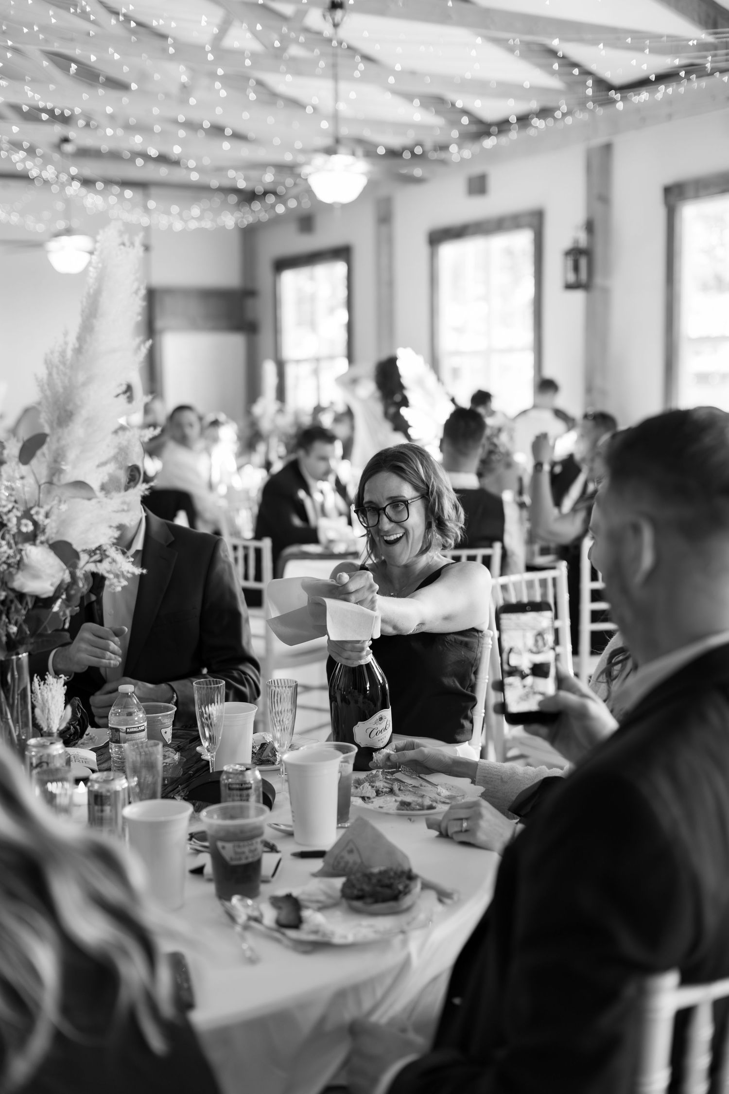 Black and white photo of wedding guest pouring champagne at elegant reception with string lights.