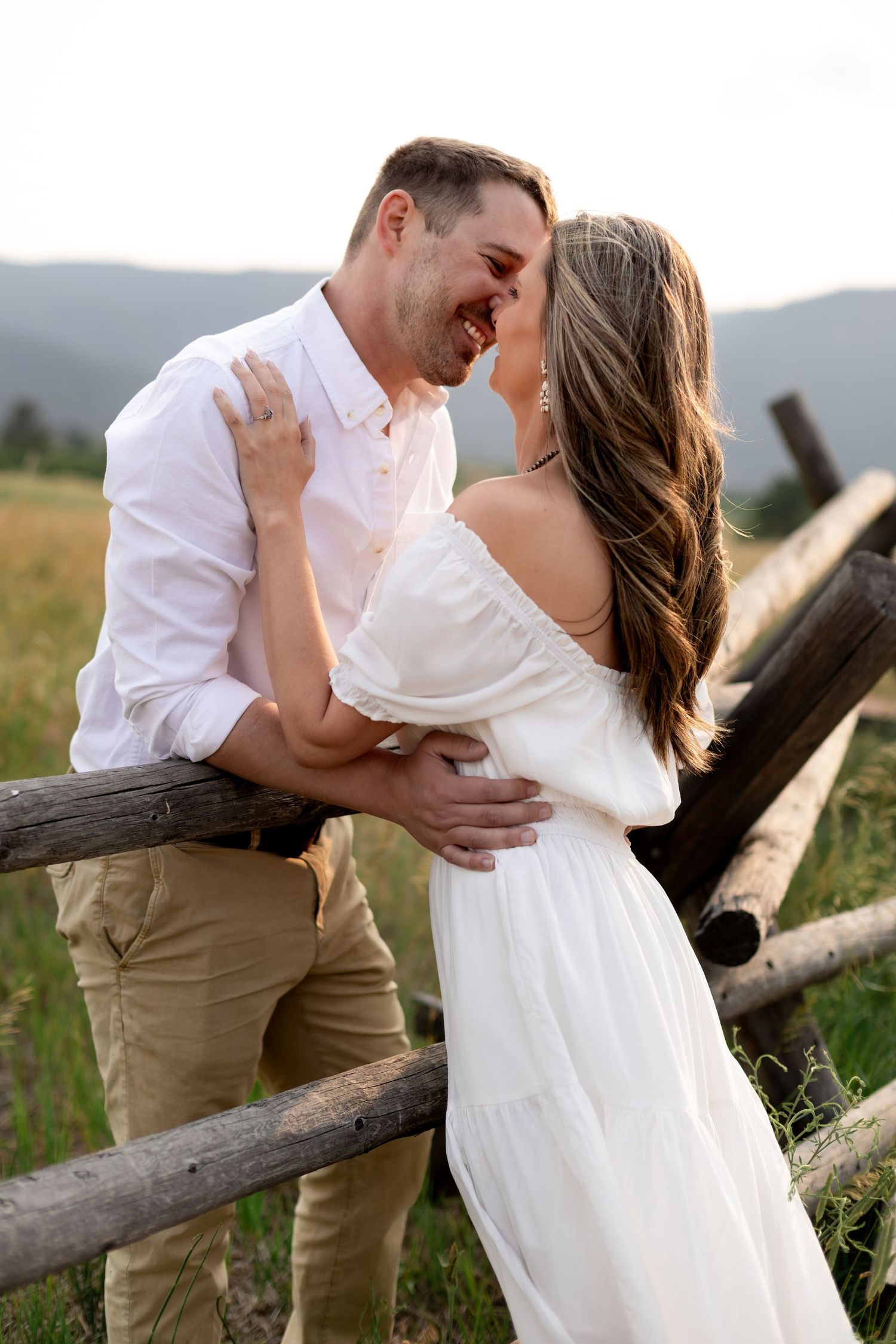 Couple sharing a romantic moment by a rustic wooden fence in a mountain meadow, woman in white dress.