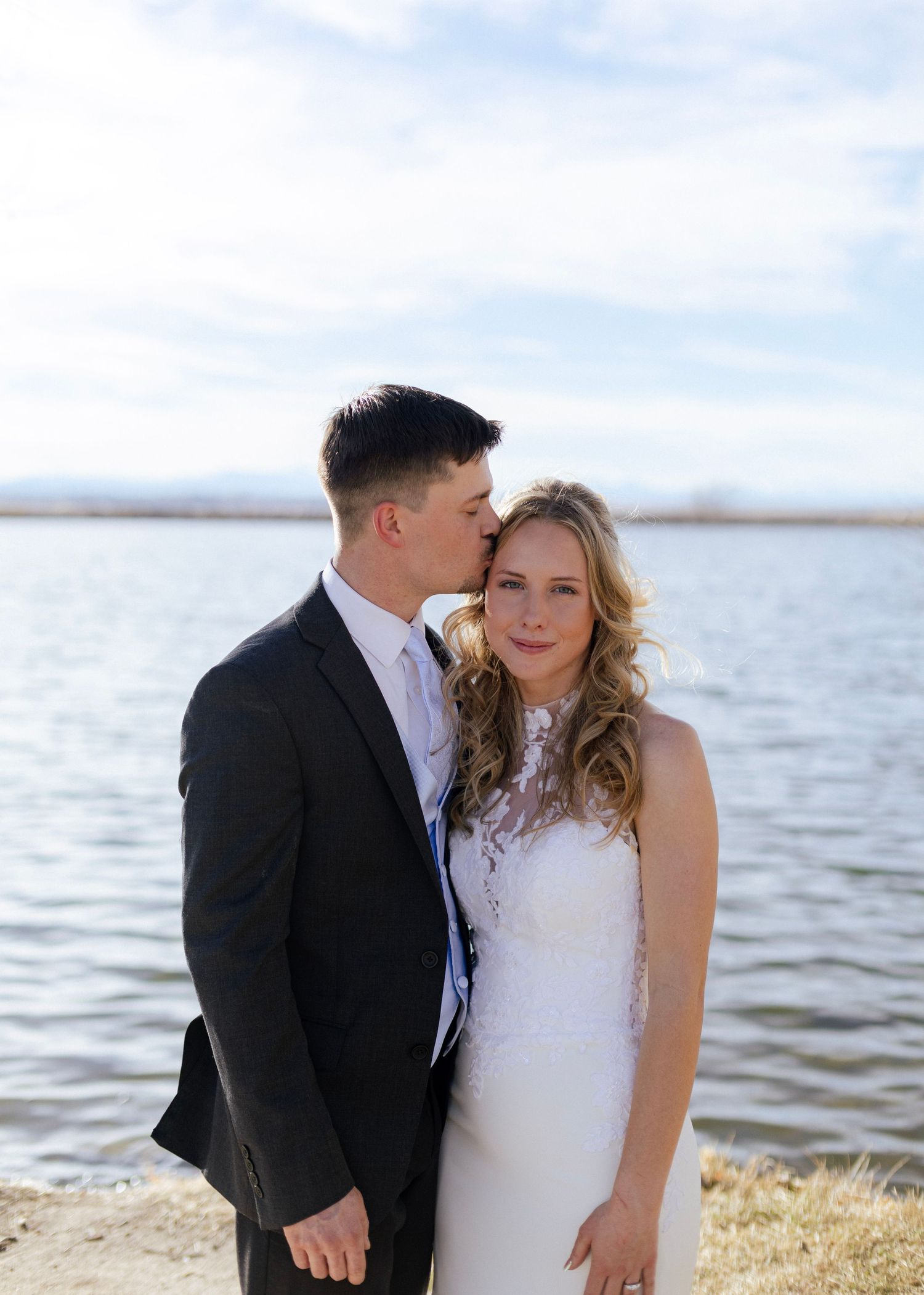 Groom gently kisses bride on the temple as they pose together in front of a serene lake backdrop.