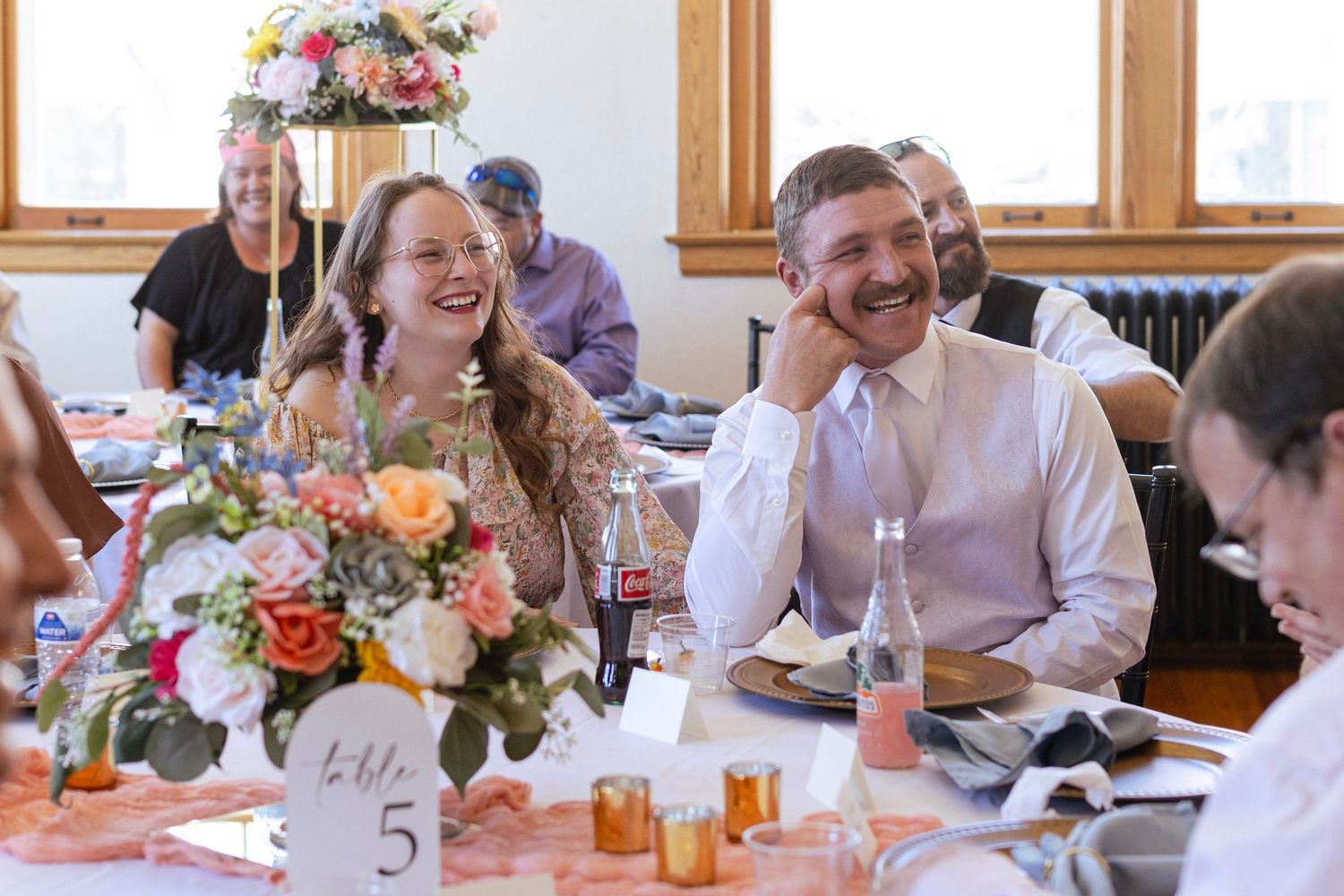 Bride and groom laughing joyfully at wedding reception table adorned with peach floral centerpieces and table number five.