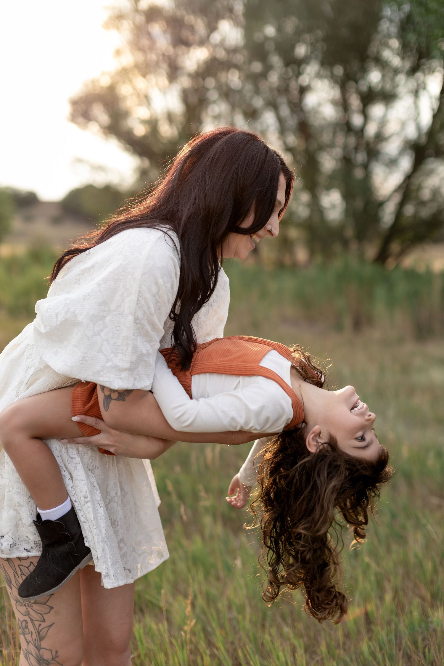 Two women laughing and playing in a field at sunset, one lifting the other upside down in a joyful embrace.