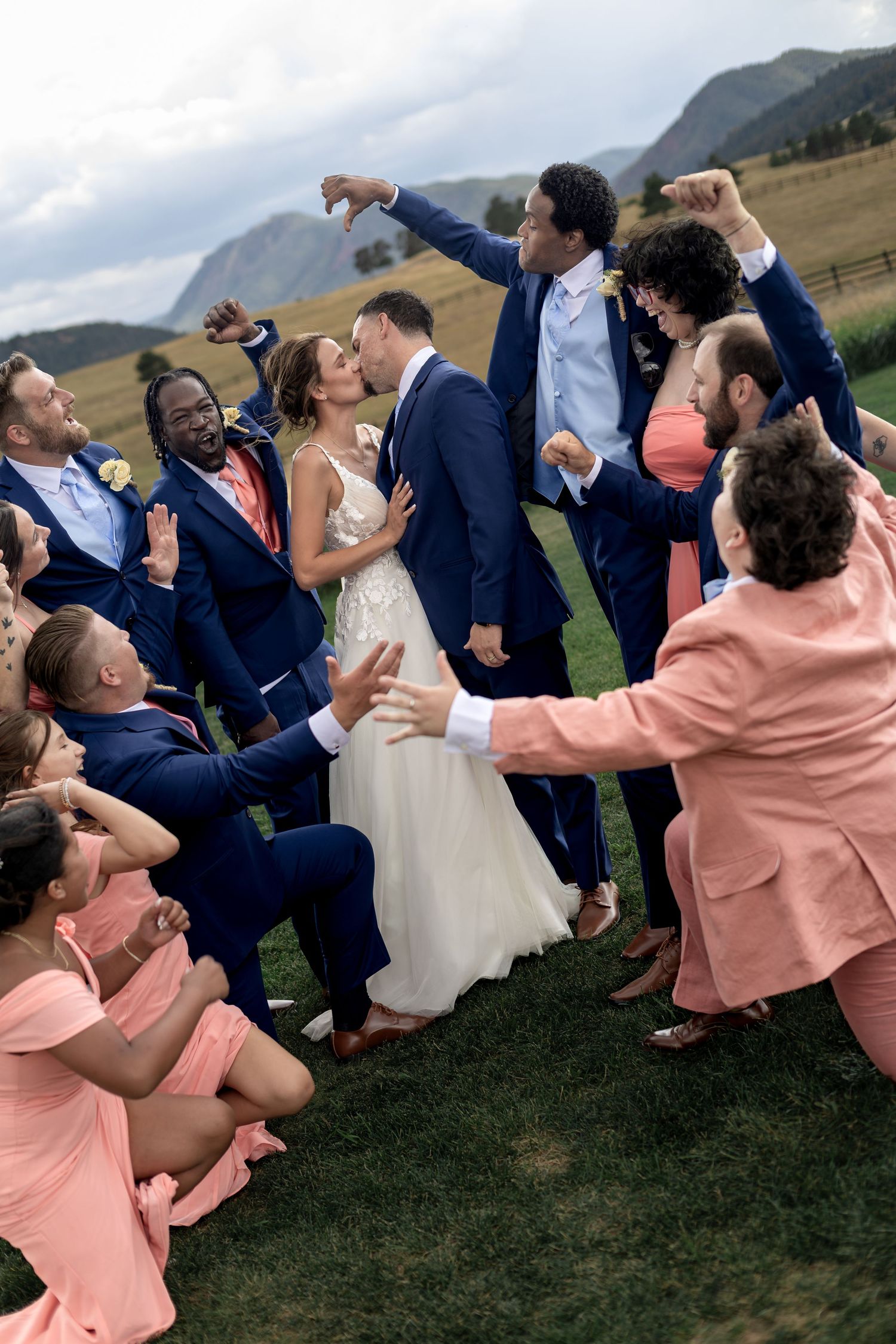 Wedding party celebrates couple kissing outdoors with mountain backdrop, bridesmaids in coral dresses and groomsmen in navy suits.