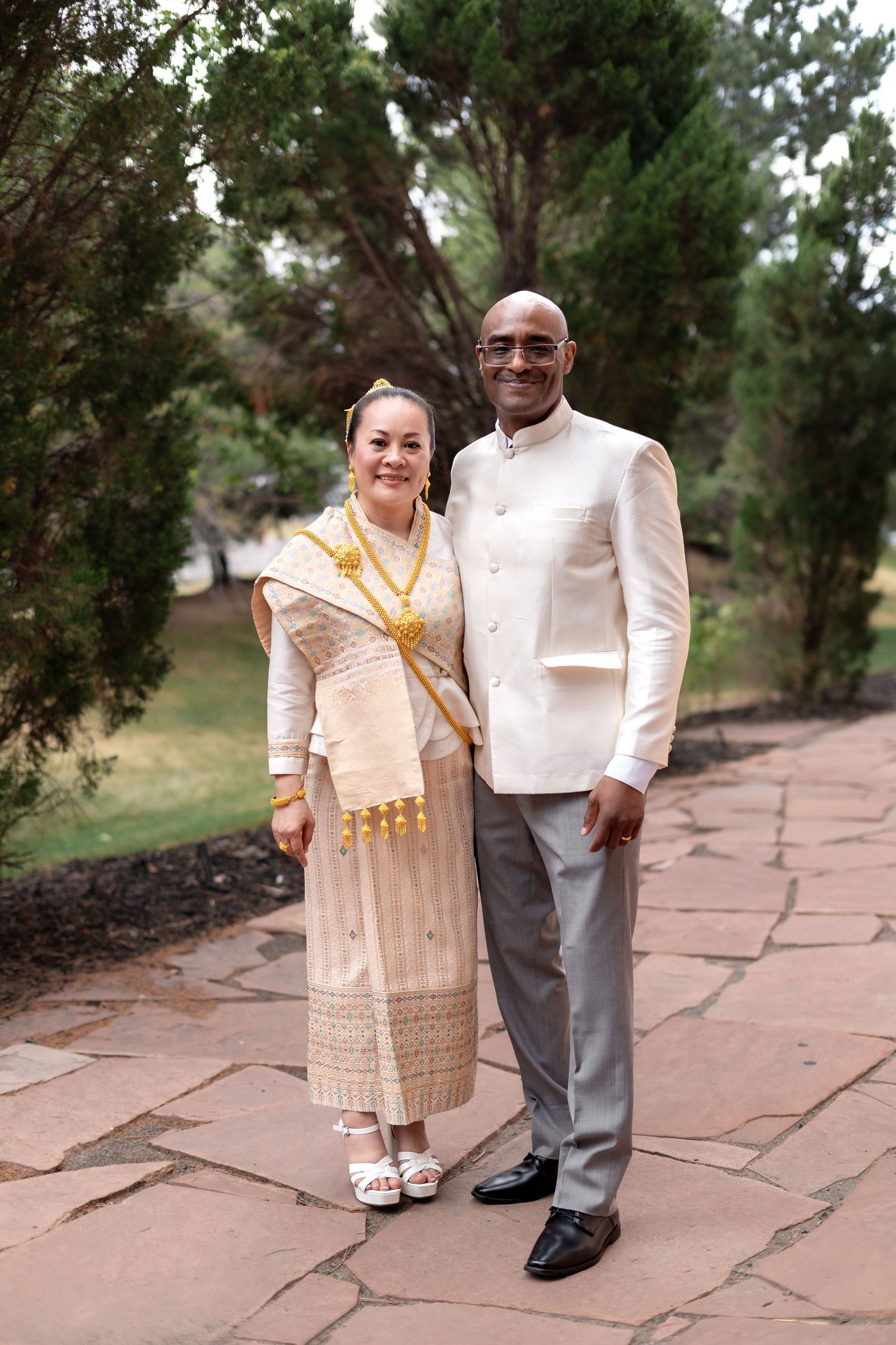 Interracial couple posing in traditional cultural wedding attire with gold jewelry on a stone path surrounded by trees.