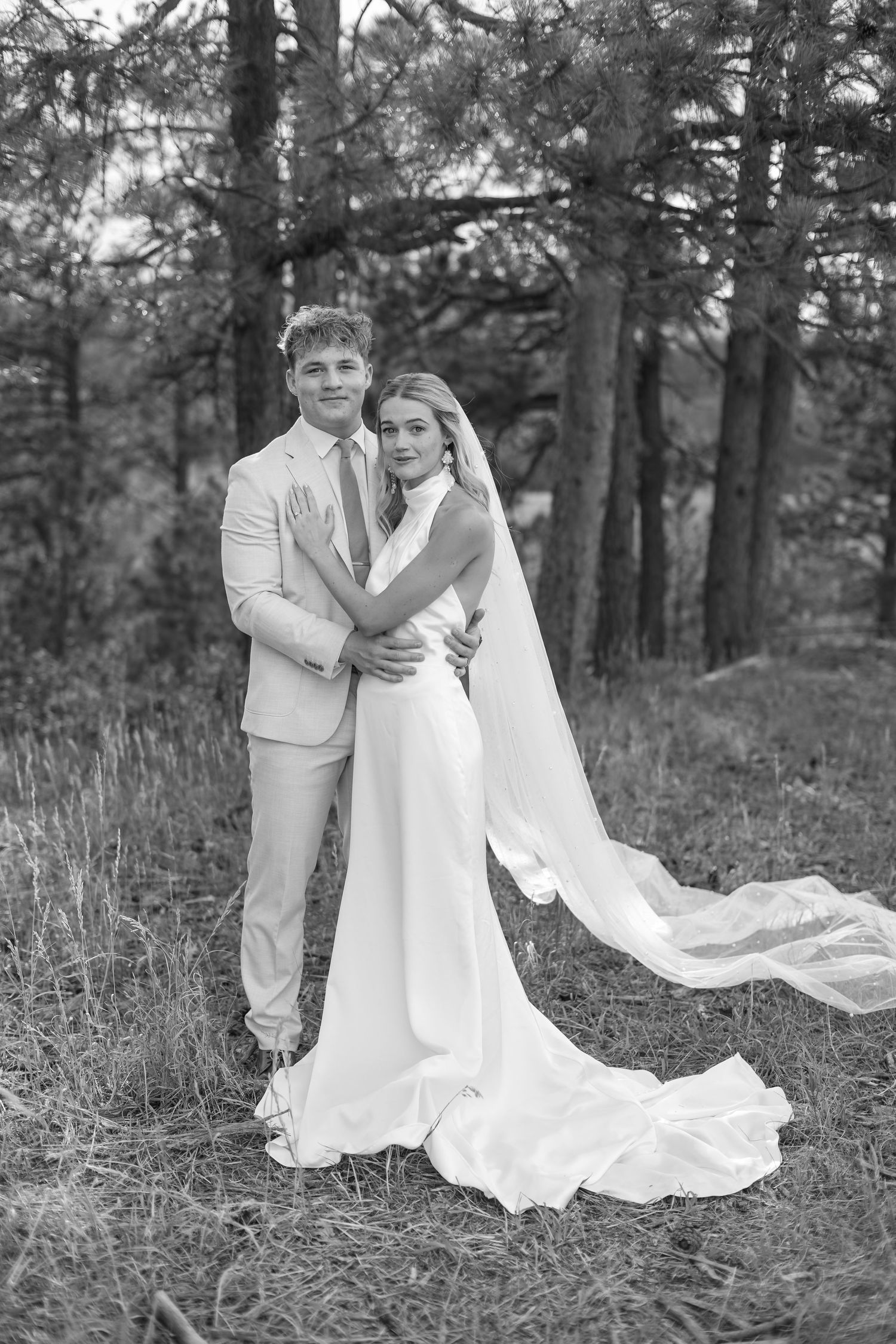 Black and white photo of bride and groom embracing in a pine forest, bride wearing flowing gown with long veil.